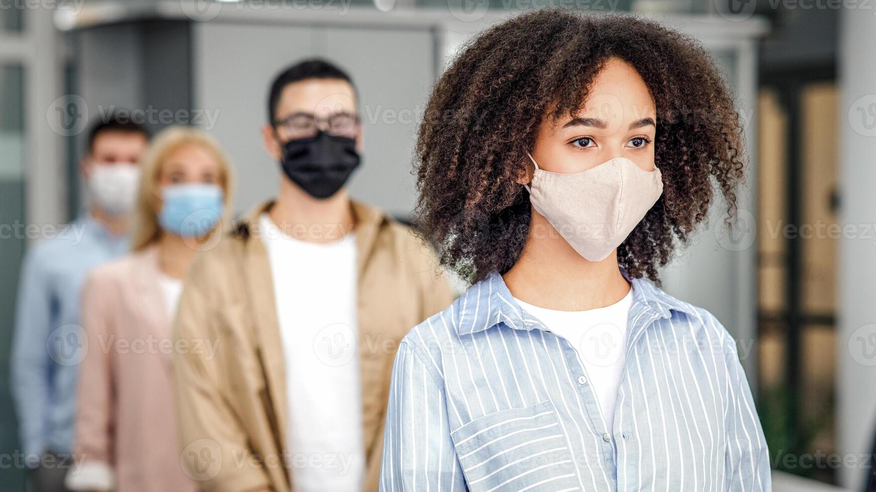 New normal and temperature check before work. Millennial multiracial employees in protective masks stand in line with social distancing at office in morning, free space, blurred, indoors photo