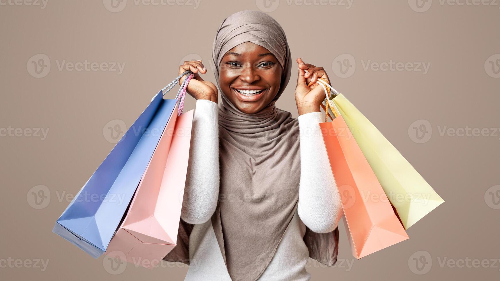 Seasonal Sales Concept. Excited black muslim woman in hijab holding lots of shopping bags, african shopahilic lady carrying purchases and smiling at camera, standing over light studio background photo