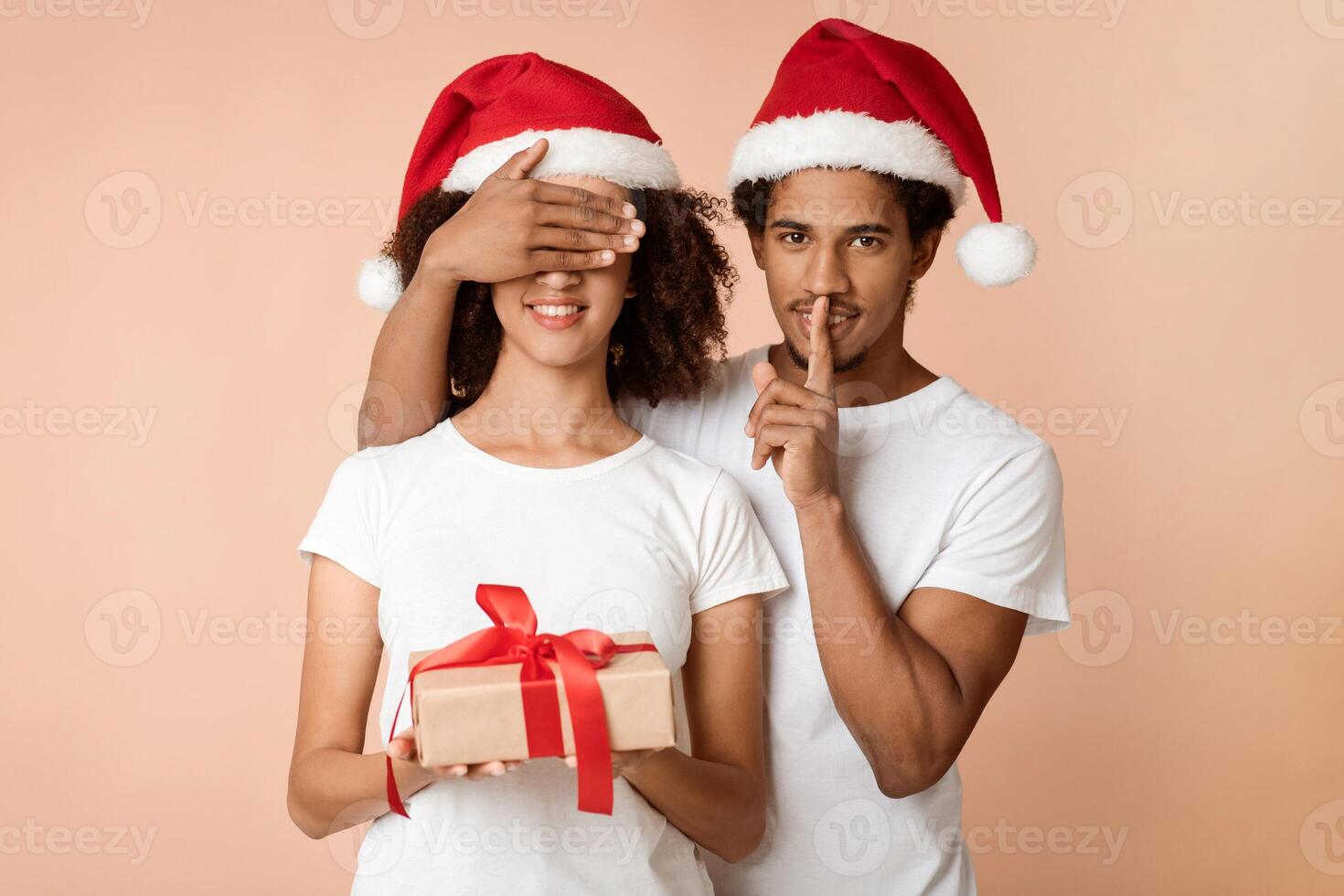 New Year presents, winter holidays. African american man in hat shows sign of silence, closes eyes to his wife and gives her box with gift isolated on light background, studio shot, free space photo