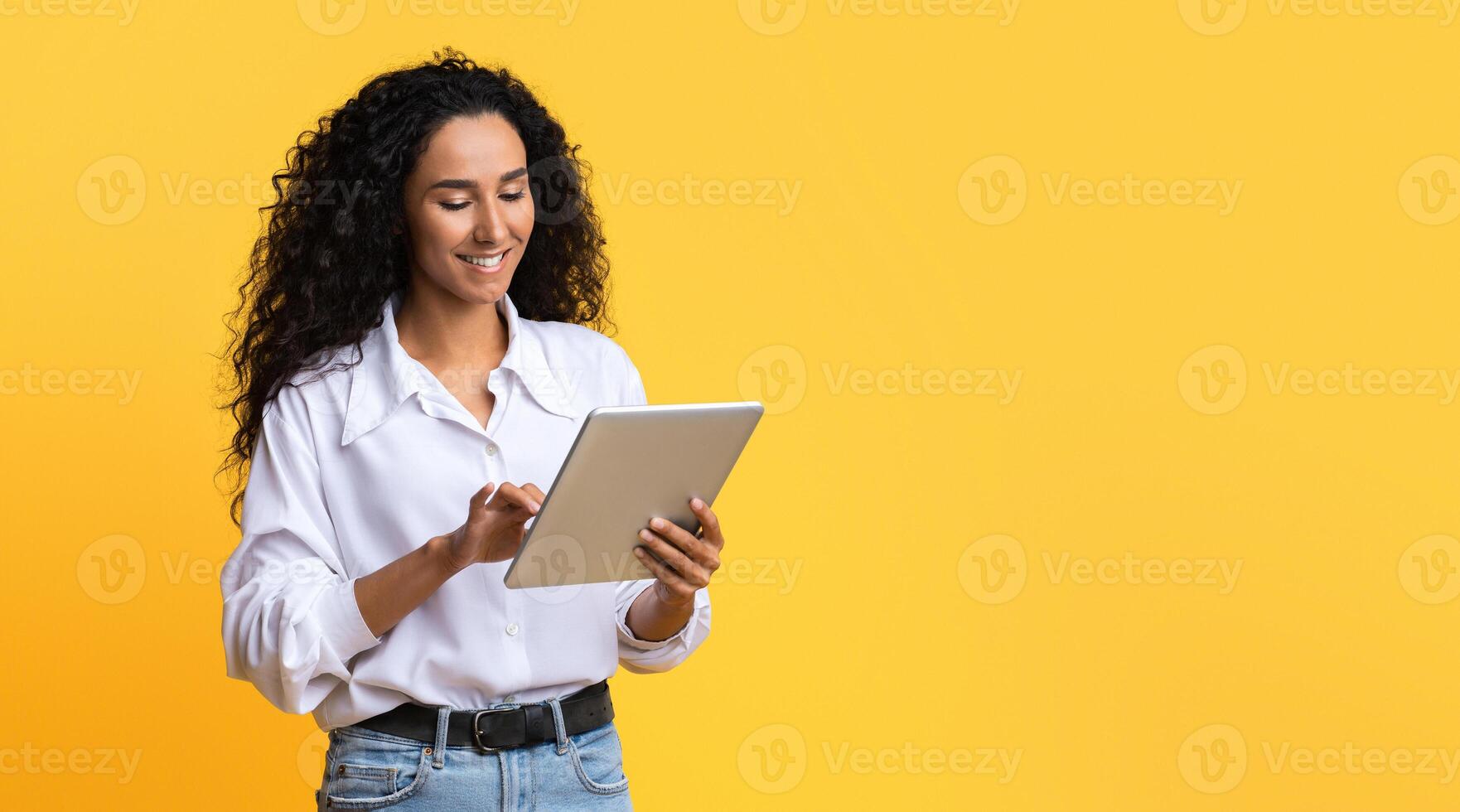 Young Brunette Woman Using Digital Tablet, Browsing Internet Or Social Networks, Standing Over Yellow Studio Background, Enjoying Modern Technologies, Checking New App Or Website, Copy Space photo