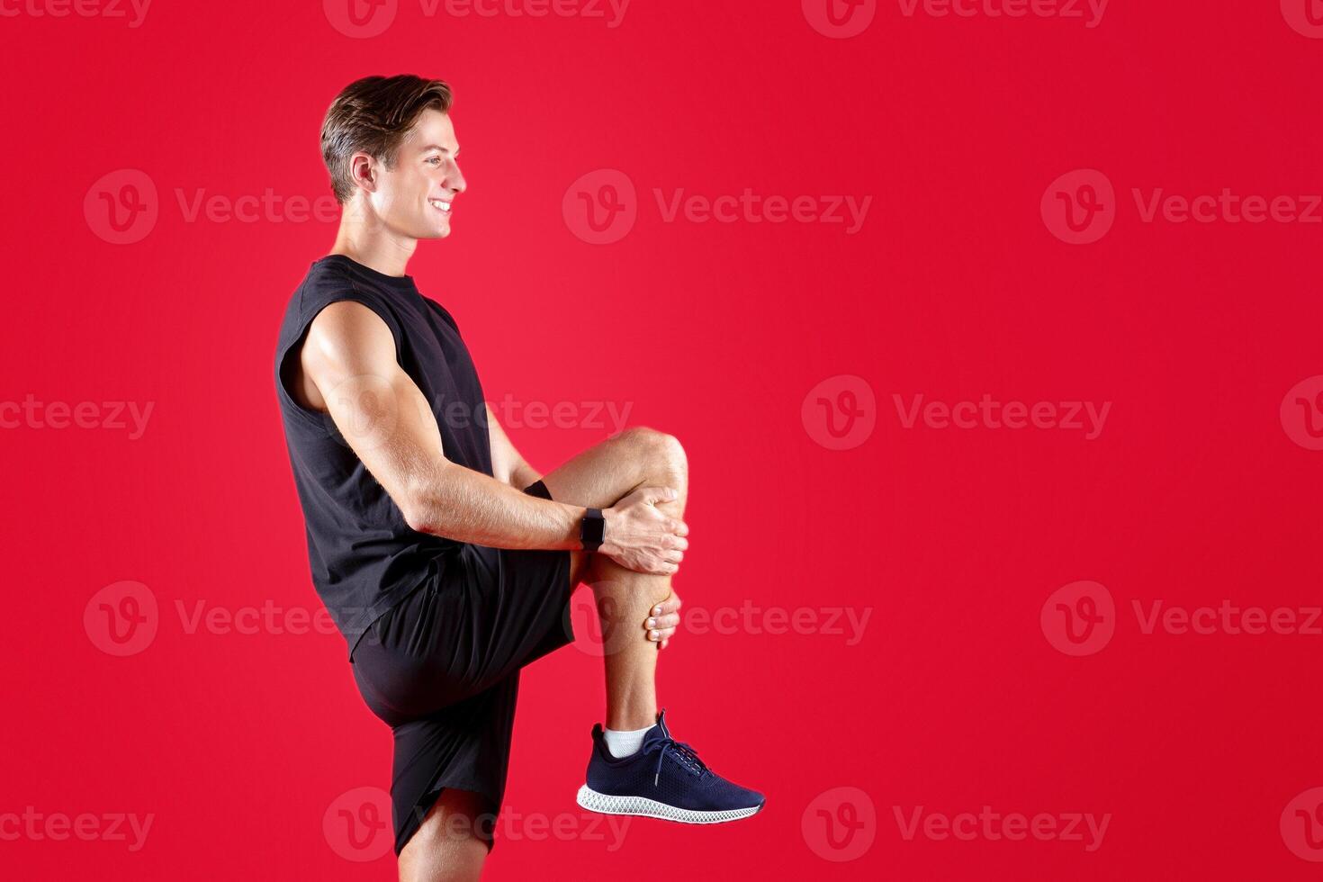 Full length portrait of sporty Caucasian man stretching his leg over red studio background. Fit millennial guy in sports clothes warming up before workout, doing flexibility exercises photo