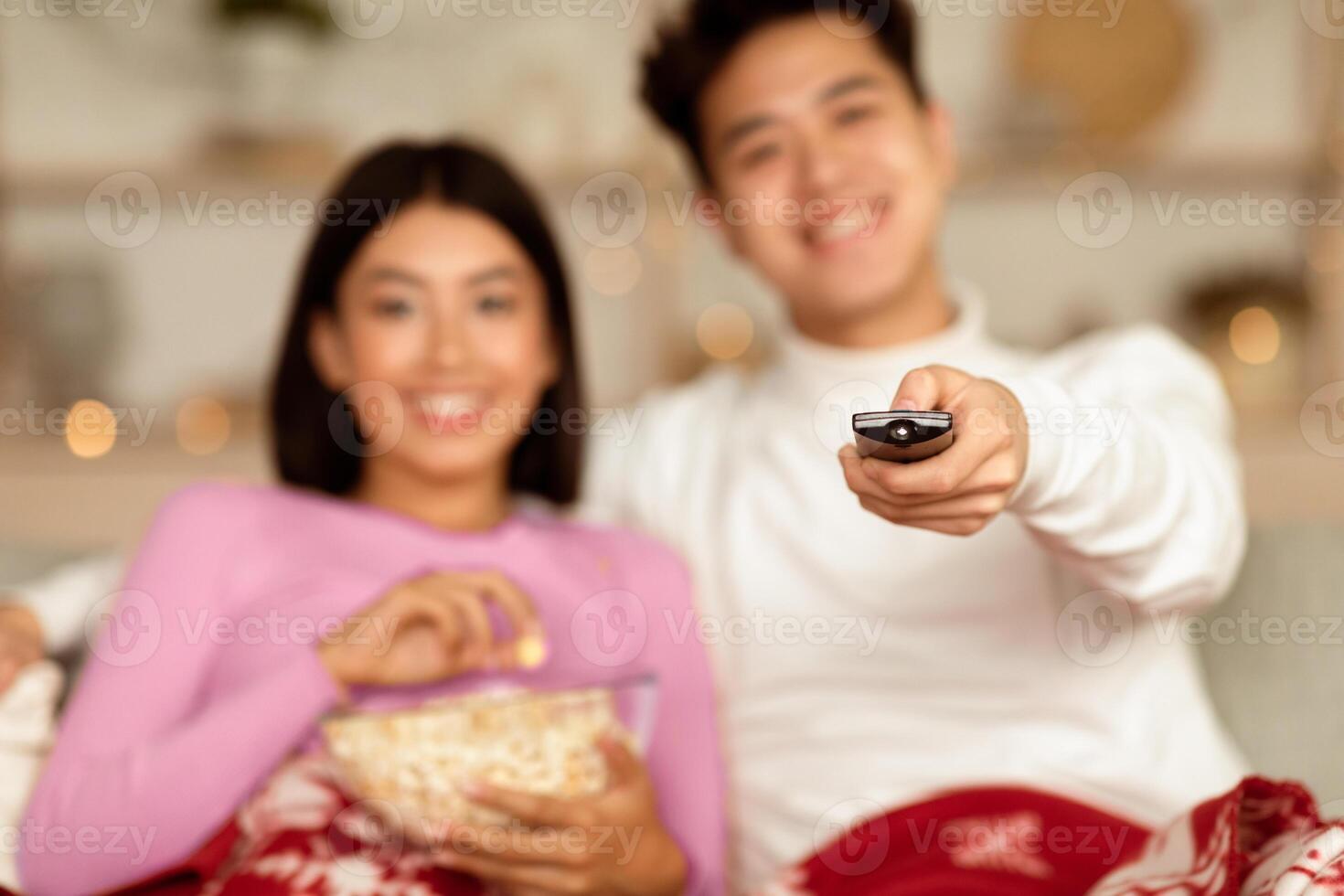 Happy Asian Couple Watching TV On Christmas, Enjoying Favorite Xmas Movies And Popcorn Sitting On Couch At Home, Focus On Remote Control. Family New Year Eve Leisure. Shallow Depth photo