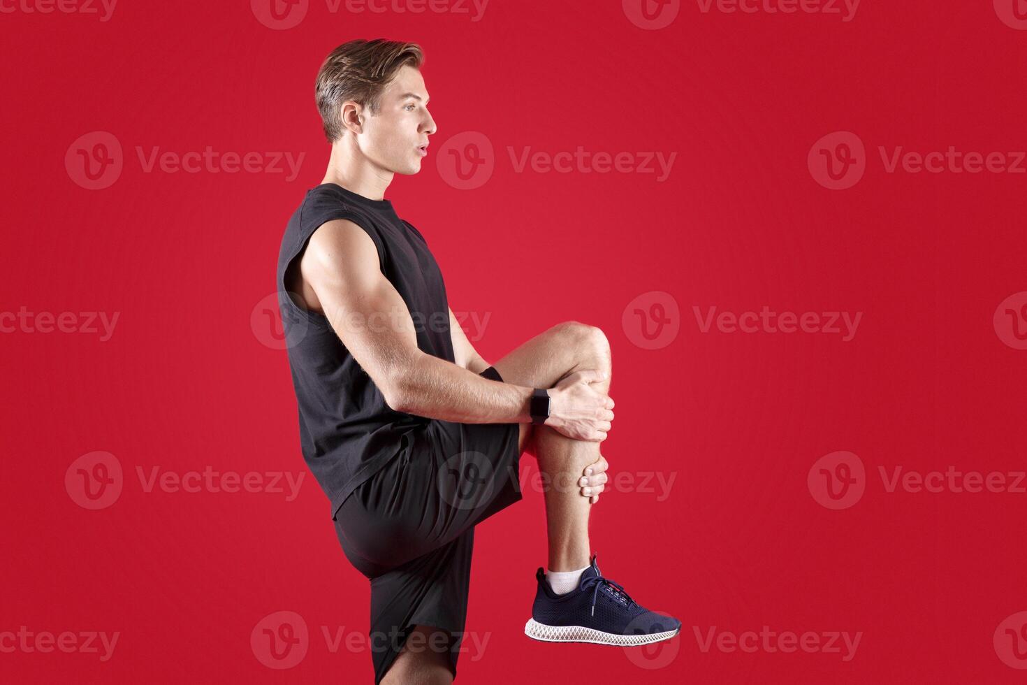 Flexible millennial man stretching his leg over red studio background, side view. Athletic young guy in sportswear warming up before training, practicing flexibility exercises photo