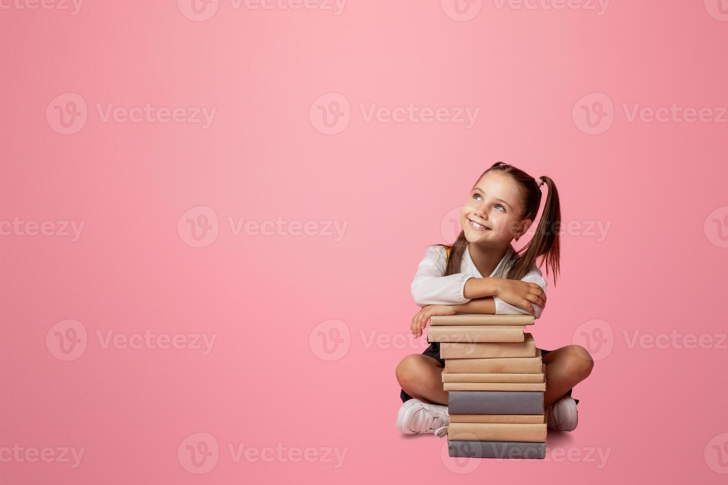 Cheerful schoolgirl dreaming, sitting at book stack and, looks at empty space. Isolated on pink background, studio shoot photo