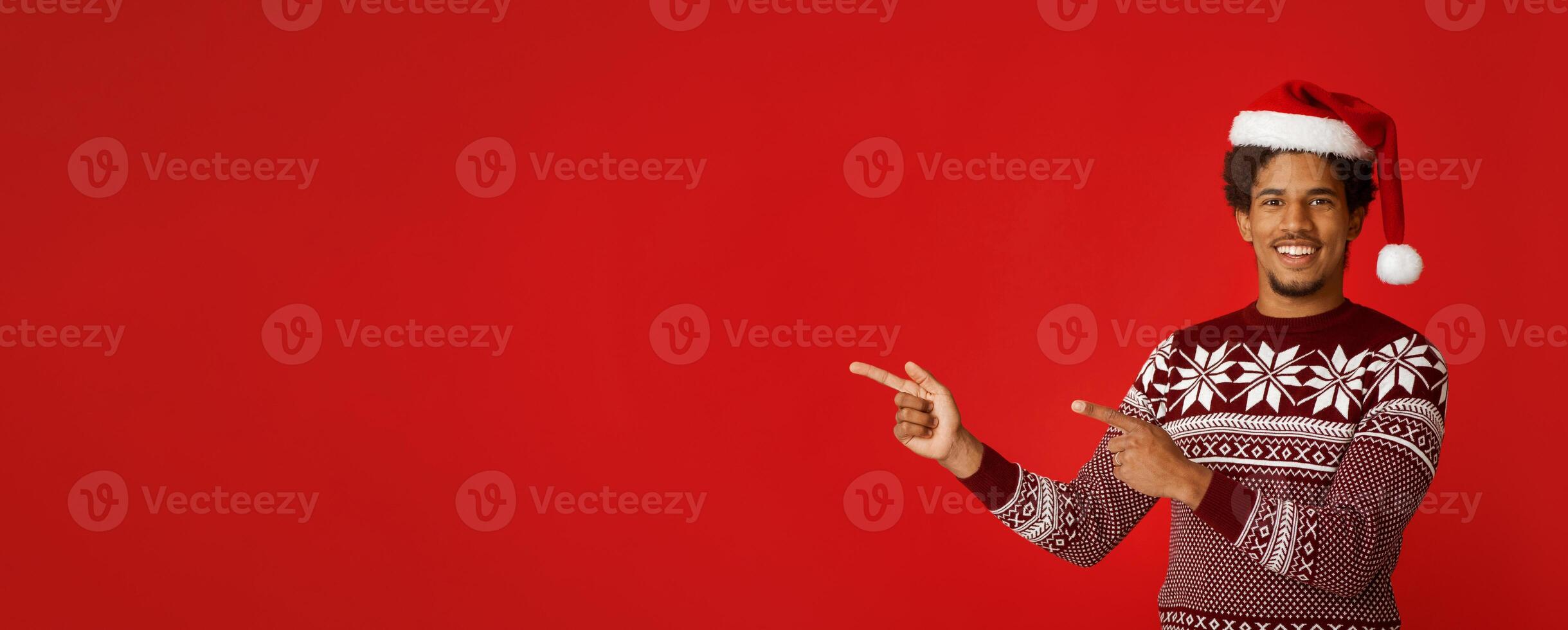 Recommendation for New Year and Christmas. Happy young african american man in santa claus hat and sweater with pattern points fingers at free space, isolated on red background, panorama, cut out photo
