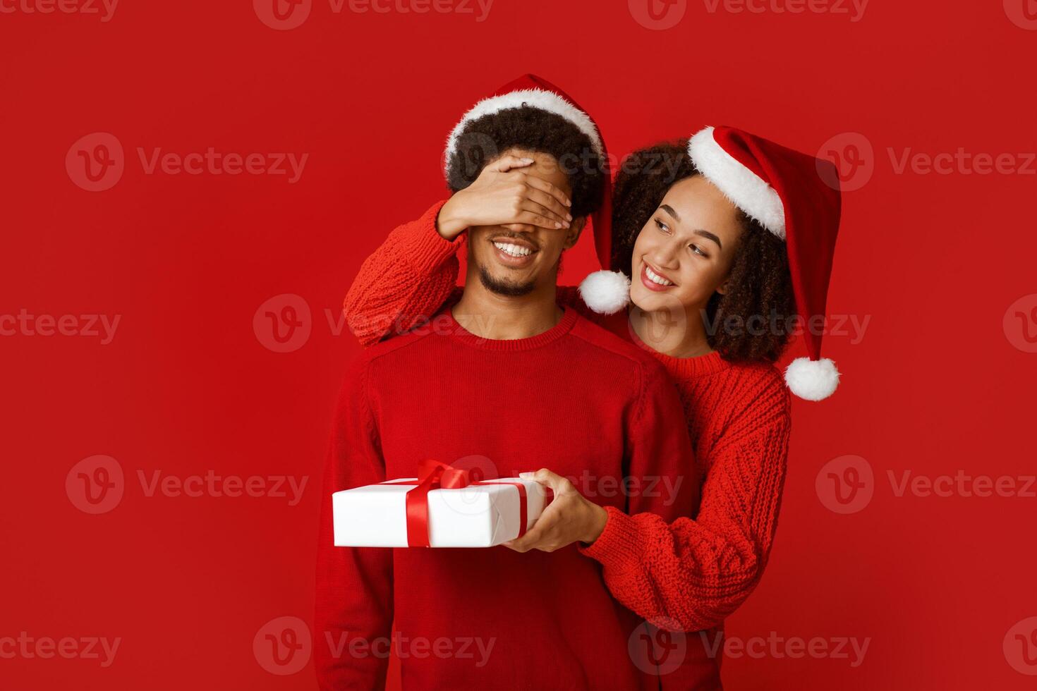 Pleasant surprise for soulmate partner and present at New Year and Xmas. Happy african american lady in Santa hat closes eyes of man in sweater and gives gift, isolated on red background, studio shot photo