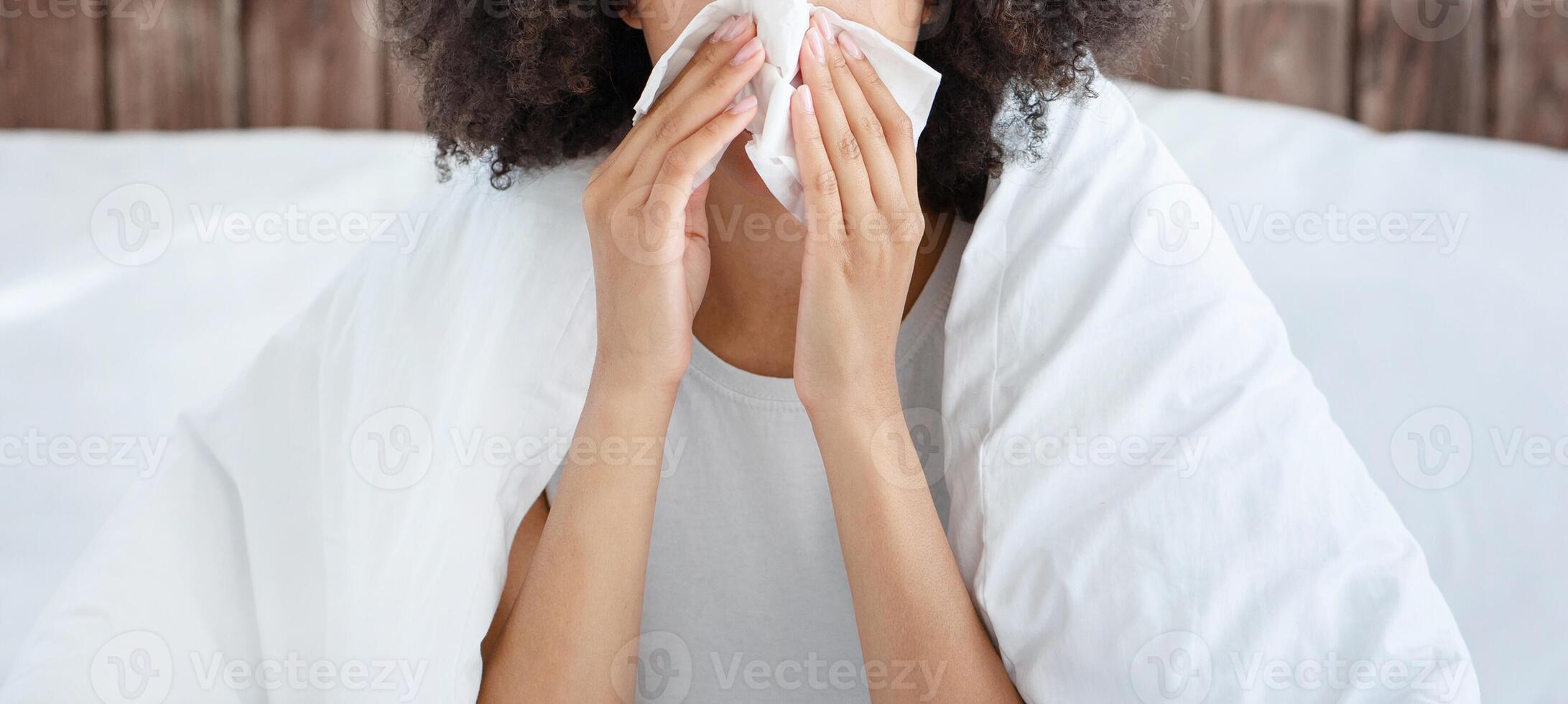 Flu and colds, illness at home and quarantine. Upset young african american lady sits on bed in blanket and wipes nose with napkin, suffering from fever in cozy bedroom interior, free space, cropped photo