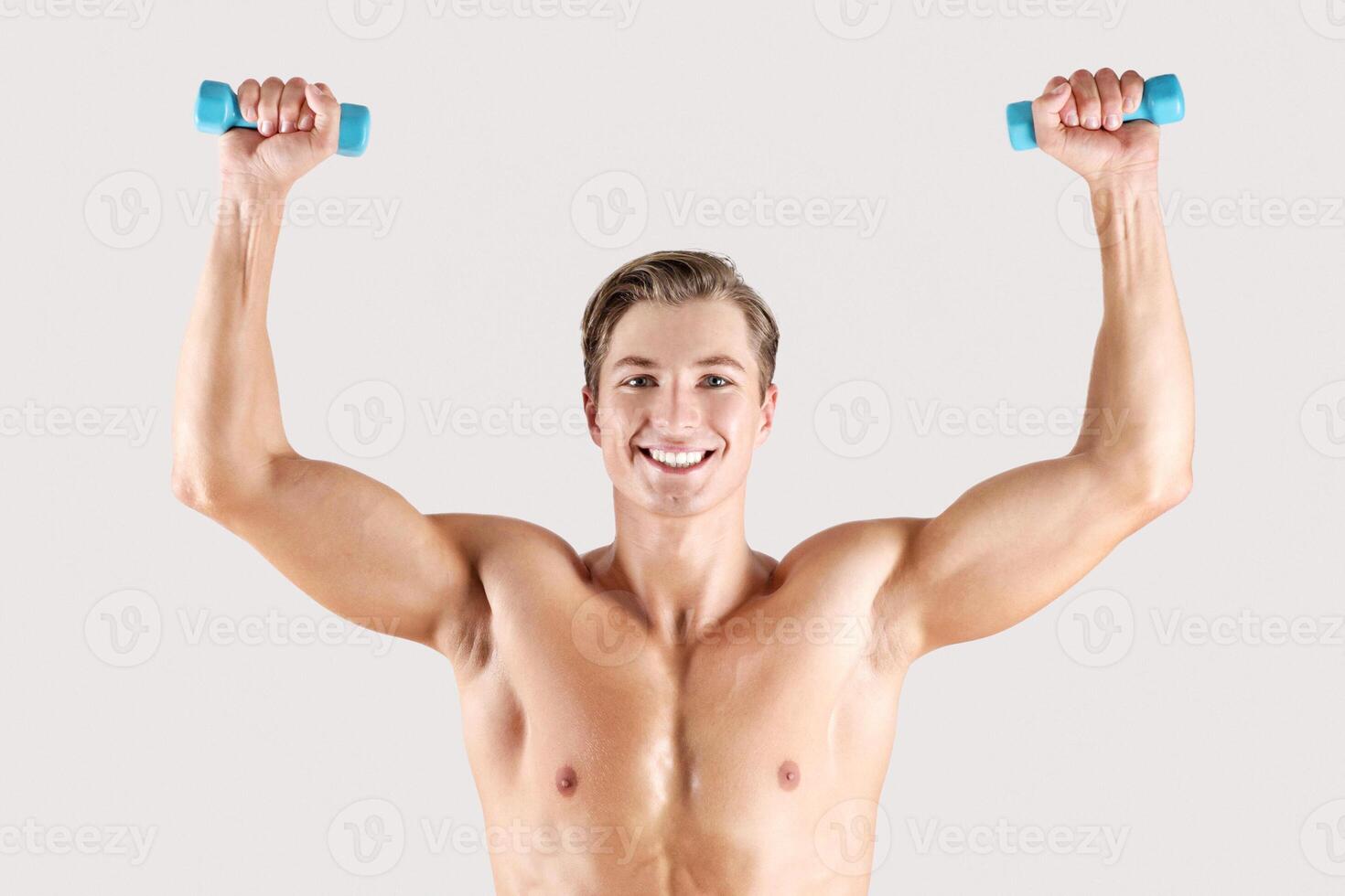 Sexy millennial sportsman lifting dumbbells, doing strength exercises on light studio background. Portrait of young guy with sexy muscular body working out with weights and smiling photo