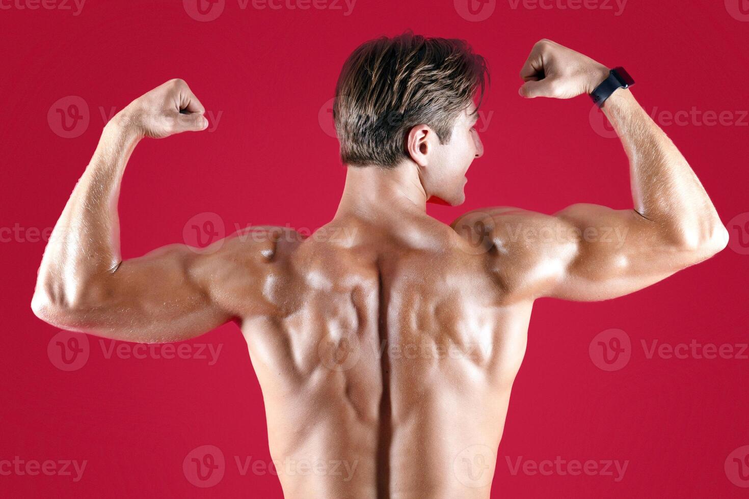 Young muscular sportsman standing with his naked back to camera and showing biceps on red studio background. Handsome athletic guy posing and demonstrating his strong muscles photo