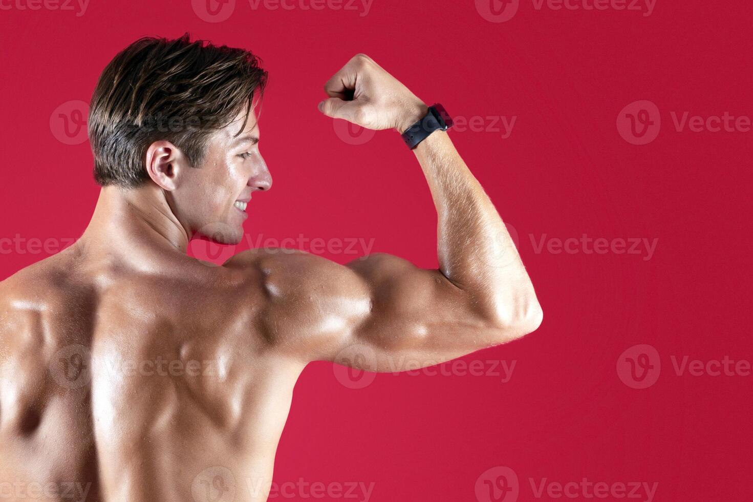Back view of happy young bodybuilder showing his biceps over red studio background. Handsome shirtless man with fit sexy body posing and smiling. Sports and fitness concept photo