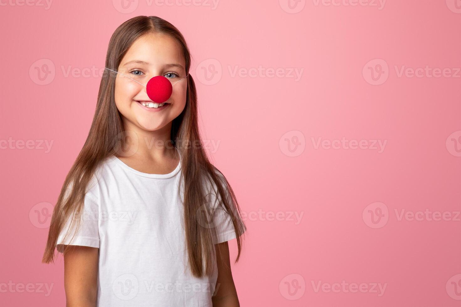 Celebrate birthday. Small girl in white t-shirt and red clown nose, isolated on pink background, studio shot photo