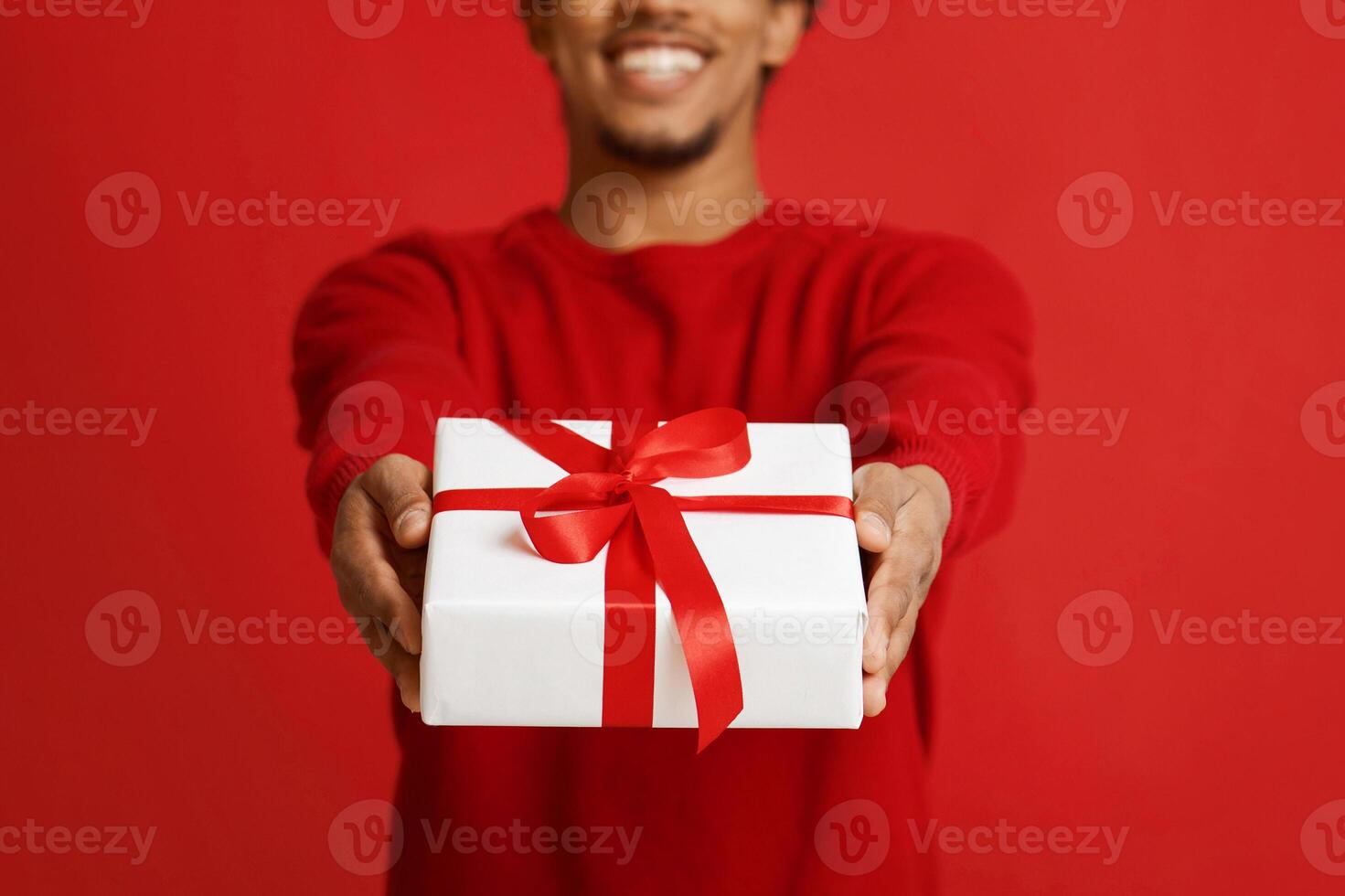 Gift box in hands wrapped in decorative paper. Happy african american young man in Santa Claus hat and sweater giving box, isolated on red background, close up, panorama, copy space, studio shot photo