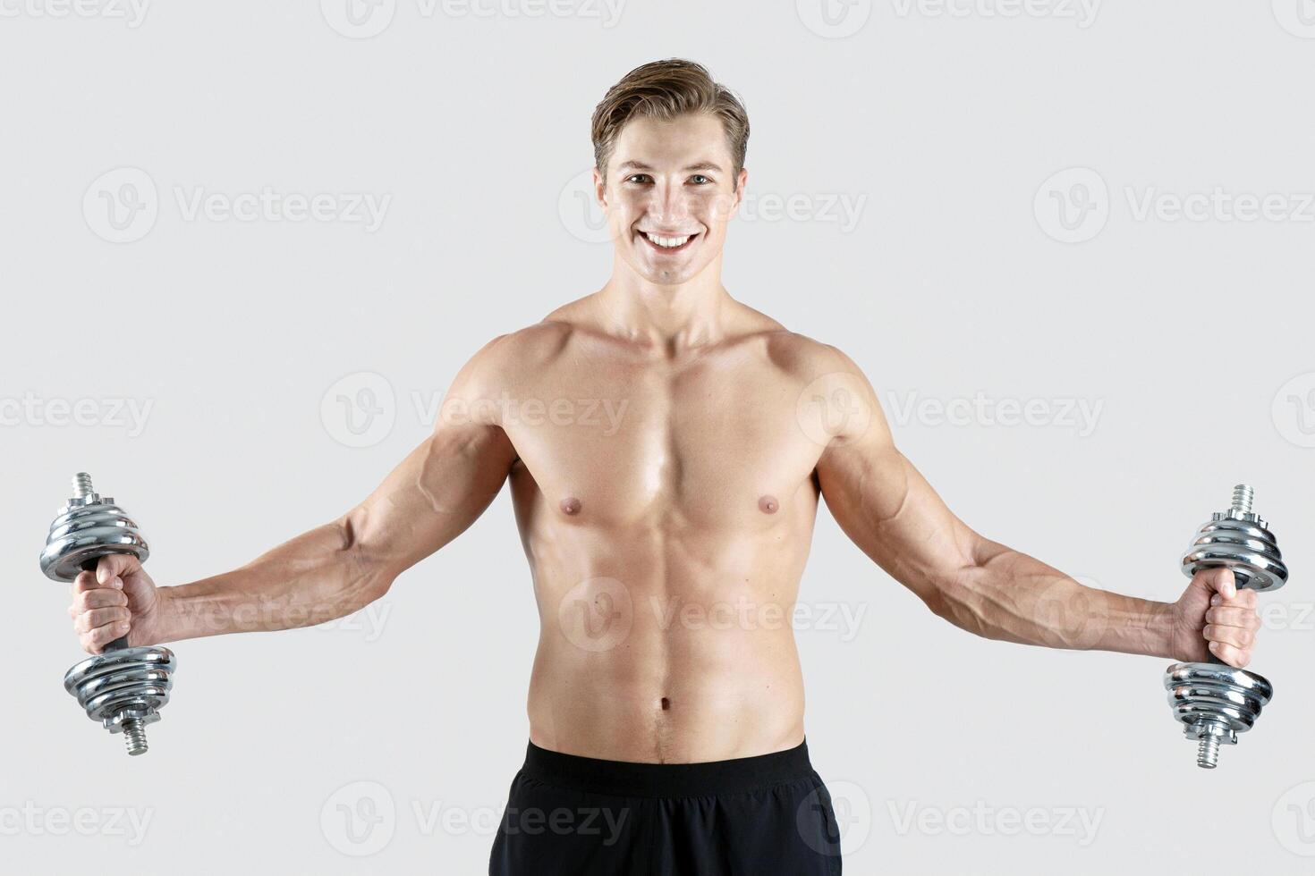 Portrait of strong young sportsman with bare chest and six pack abs working out with dumbbells on light studio background. Muscular millennial guy with sexy body lifting weights, pumping muscles photo