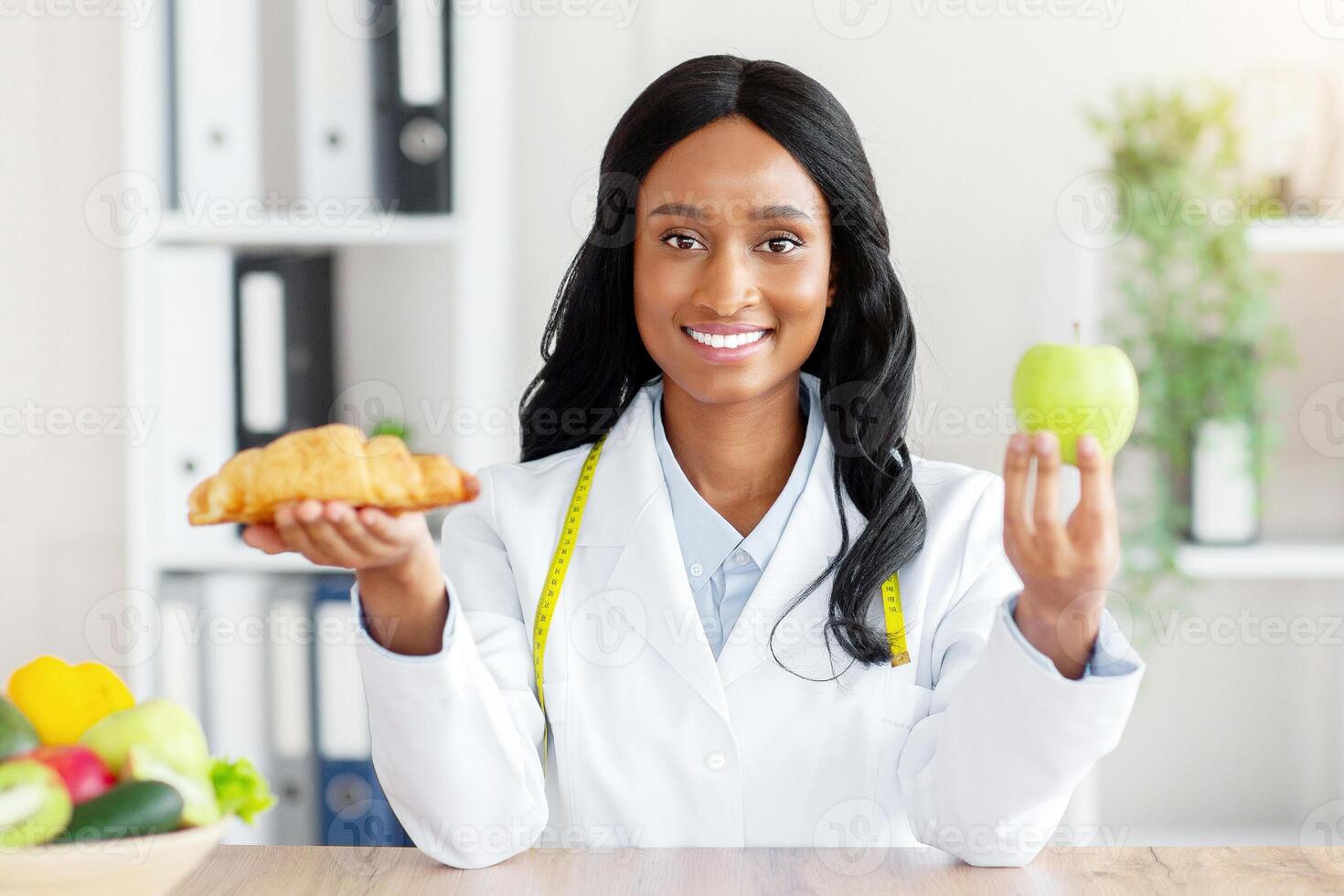 Making healthy food choices. Beautiful female nutritionist holding croissant and apple at table in medical office. African American dietitian selecting between healthy and junk products photo