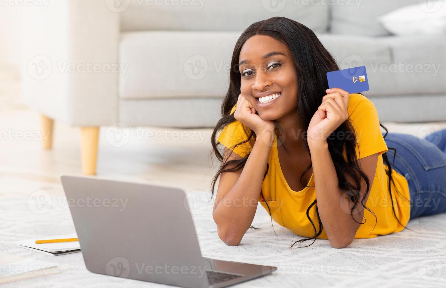 Online shopping. Positive black lady with laptop and credit card lying on floor at home, purchasing goods in web store. Positive young woman buying things on internet, using contactless payment photo