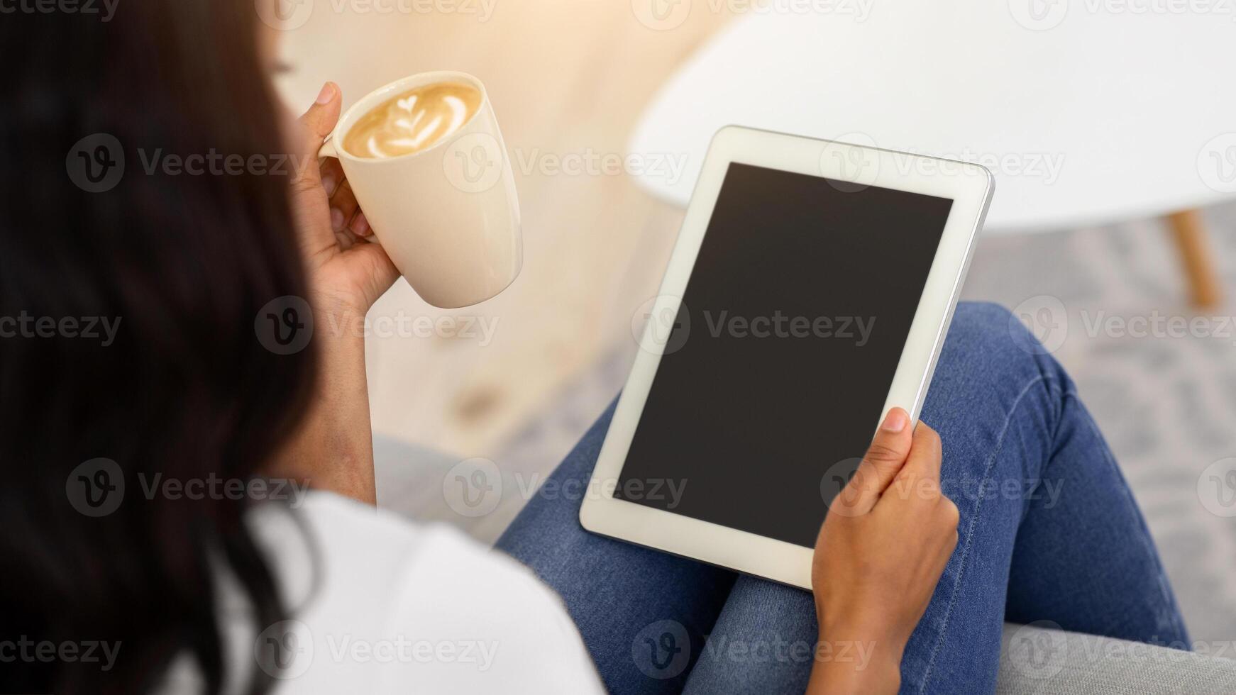 Young black woman with coffee sitting on sofa and using tablet computer with blank screen at home, mockup for your website design. African American lady working online, selective focus photo
