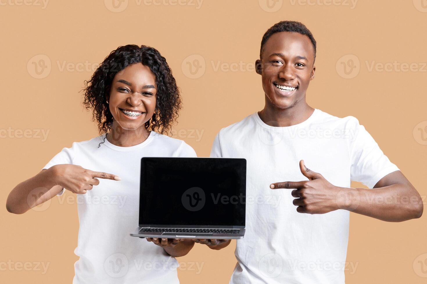 Young emotional african-american couple pointing fingers at blank black laptop screen, yellow studio background. Smiling black man and woman showing website on their computer, mockup photo