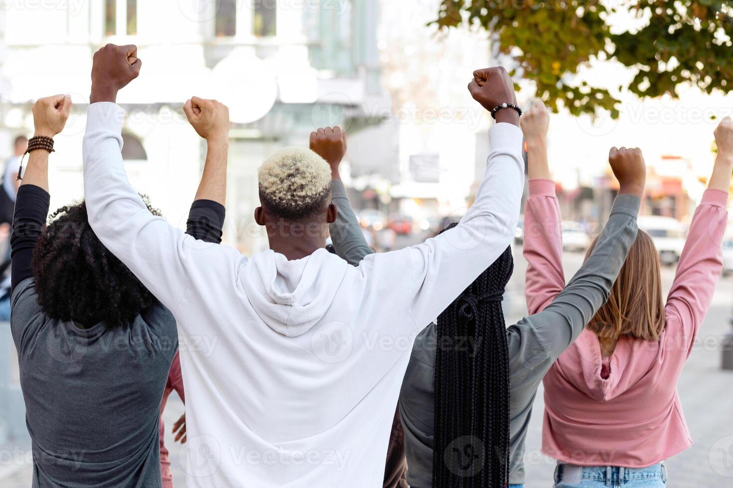 Back view of multiracial millennial demonstrators men and women in casual standing on the street, raising fists up, fighting fot human rights, defying lockdown restrictions in front of government hall photo