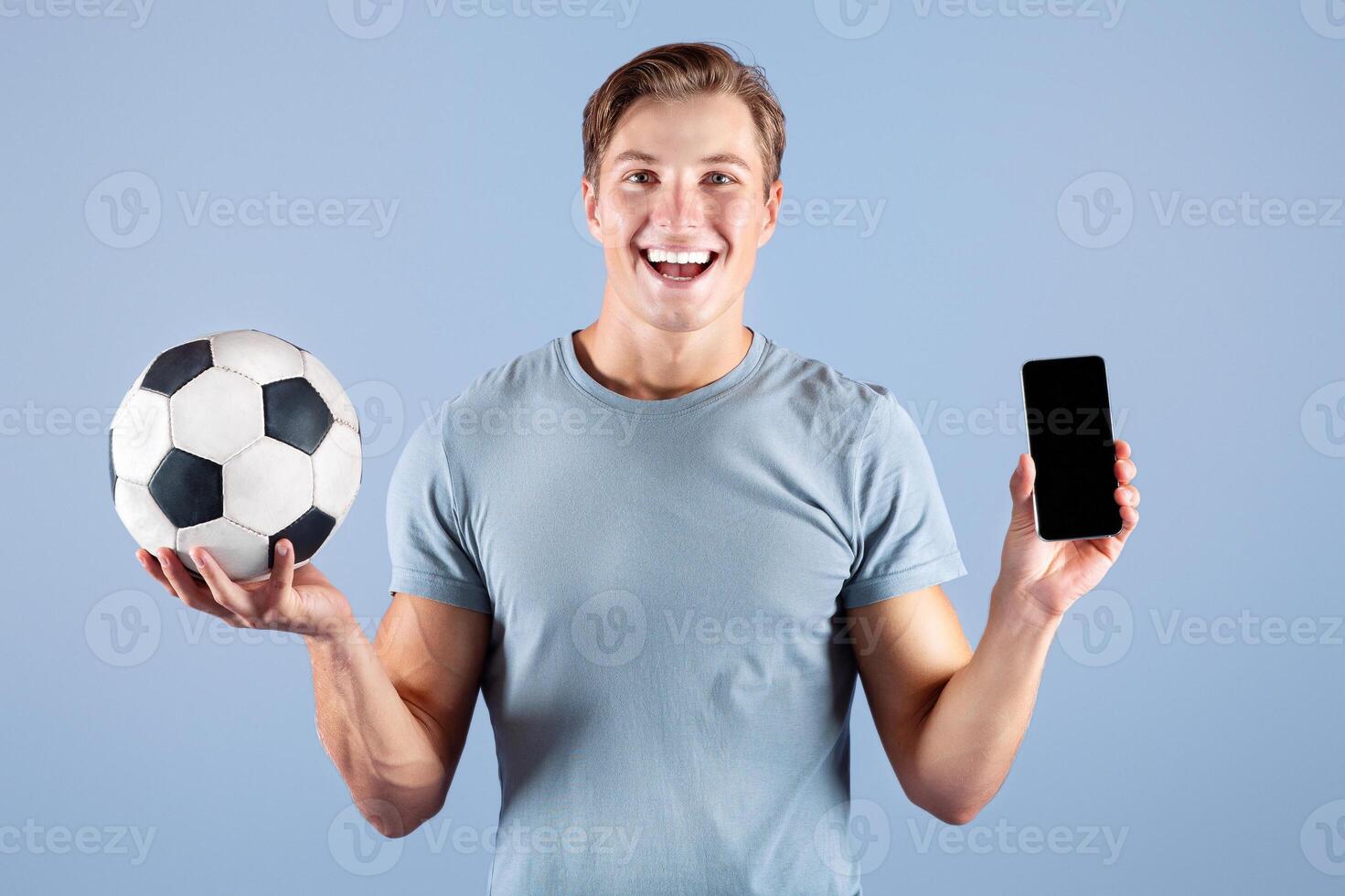 Happy young guy holding soccer ball and smartphone with blank screen on lilac studio background, mockup for design. Positive football fan showing mobile device with space for website or app photo