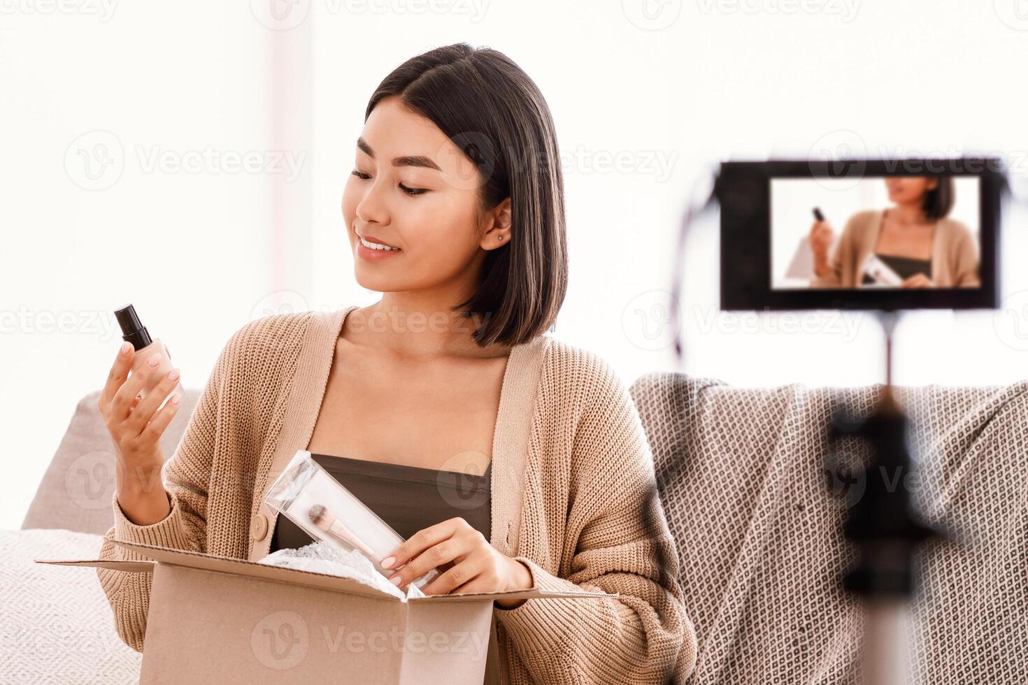 Product Review. Satisfied asian young woman vlogger holding foundation and brush, unpacking cardboard beauty box with cosmetics products, recording for her social media blog on camera at home photo