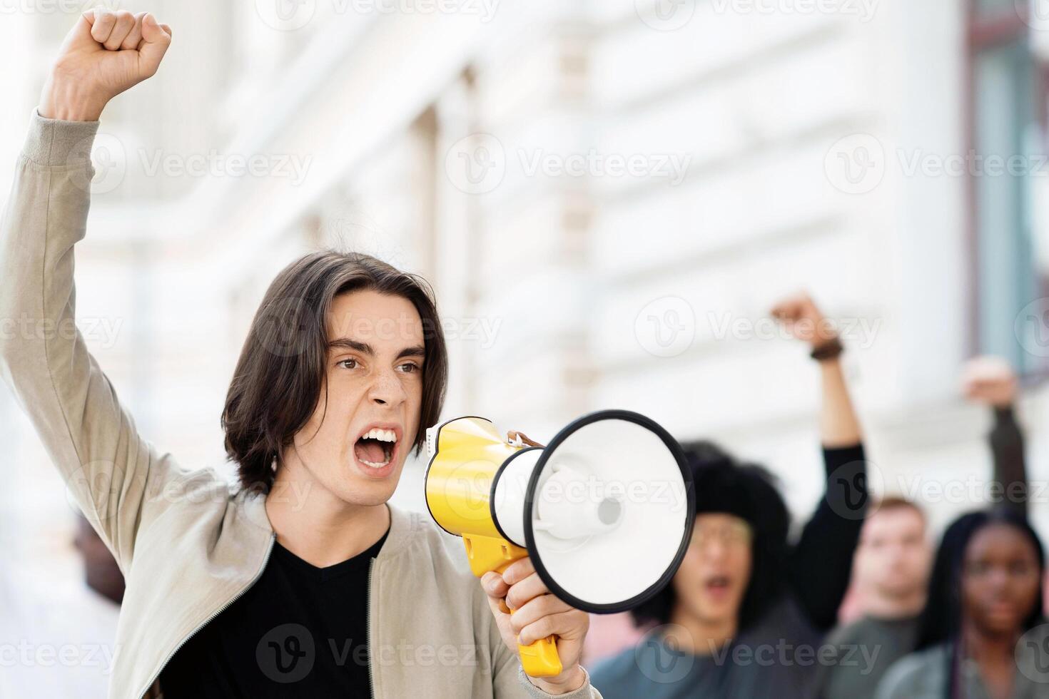 Leader of strikers young motivated man with loudspeaker yelling demands, raising fist up, leading international group of demonstrators fighting for human rights, against racism, for climate change photo