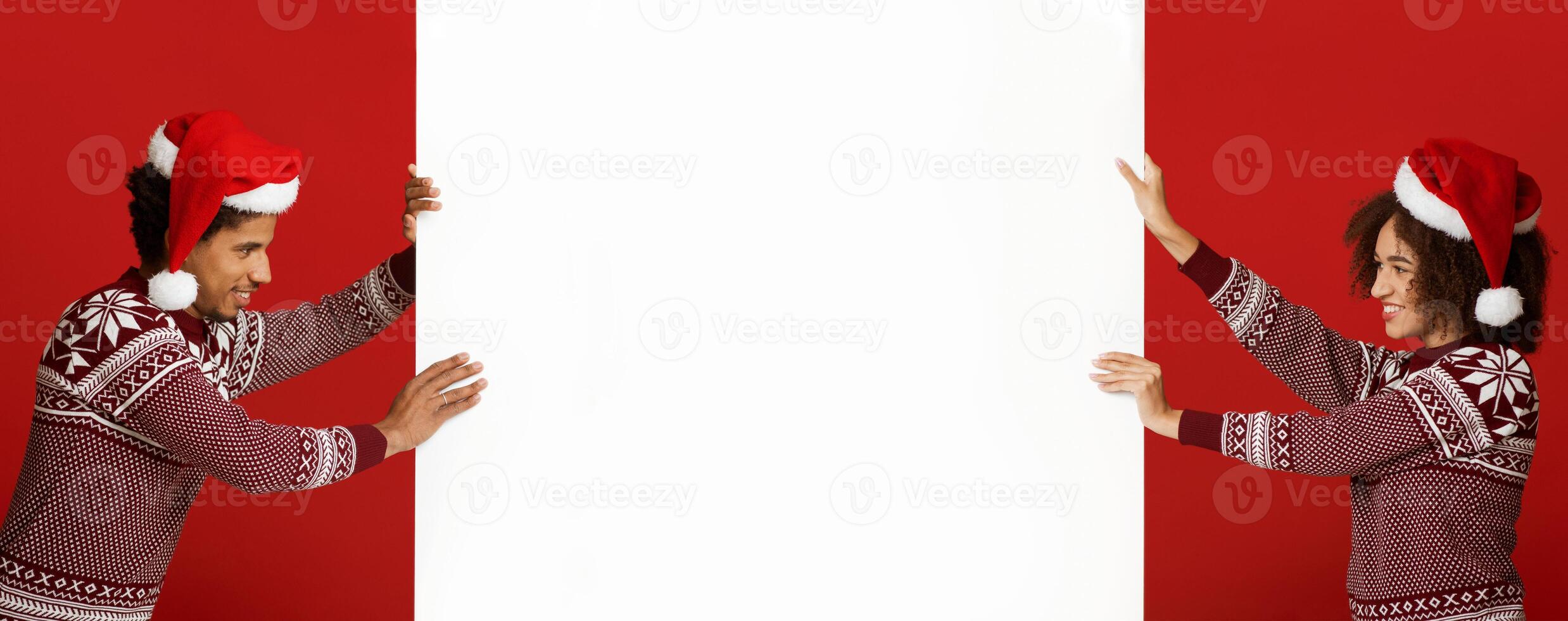 Winter, confrontation and Xmas mood. Cheerful young african american couple in Xmas hats and sweaters, push white billboard with empty space for advert isolated on red background, panorama, side view photo
