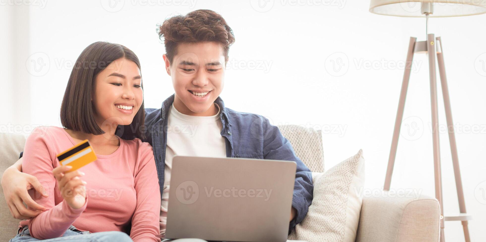 Asian Couple With Laptop Shopping Online, Holding Credit Card Paying For Order In Internet Shop Sitting On Sofa At Home. Happy Web Store Customers Buying And Purchasing Things On Weekend. Copy Space photo