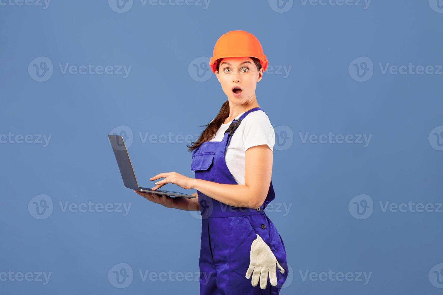 Cool Website. Shocked Handywoman In Hard Hat And Coveralls Holding Laptop And Looking At Camera With Excitement, Repair Worker Lady Using Computer For Online Shopping, Blue Background, Copy Space photo