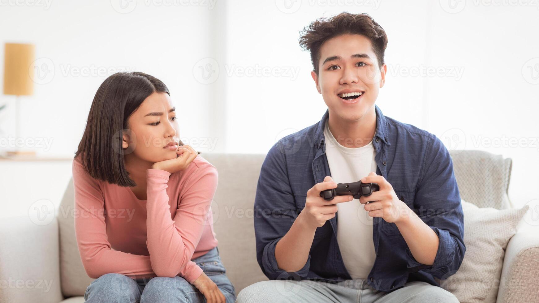 Gaming Addiction Problem. Discontented Asian Wife Looking At Infantile Husband Playing Game Sitting On Couch At Home. Computer Games, Waste Of Time, Male Emotional Immaturity Concept photo