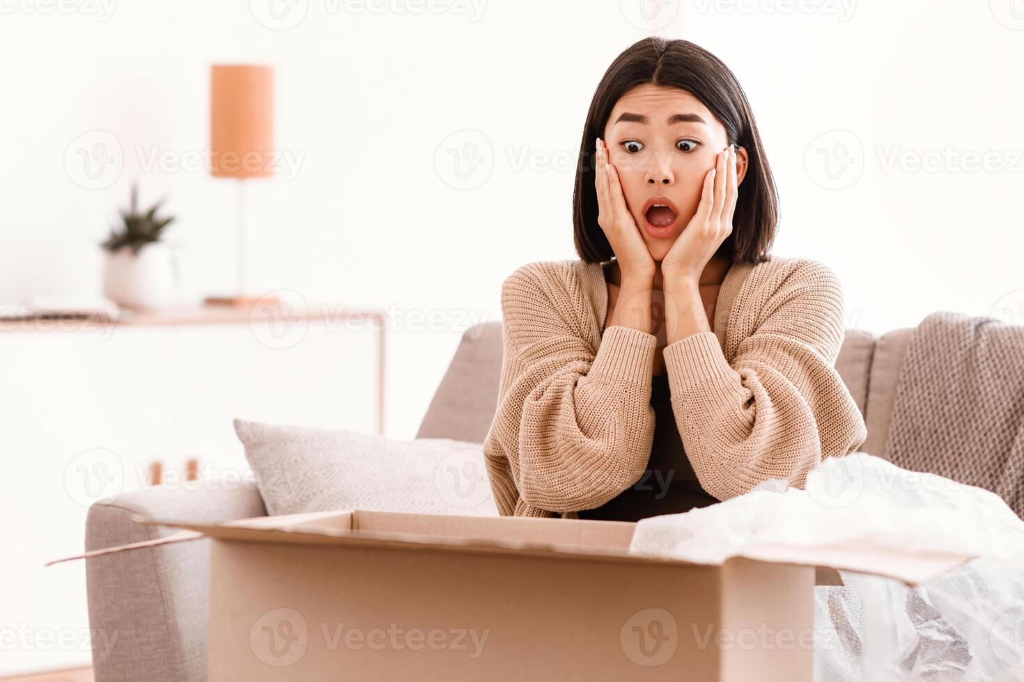 Wrong Parcel Concept. Surprised shocked asian lady touching face and screaming after unpacking cardboard box, received damaged item, sitting on the couch, feeling upset with bad wrong delivery photo