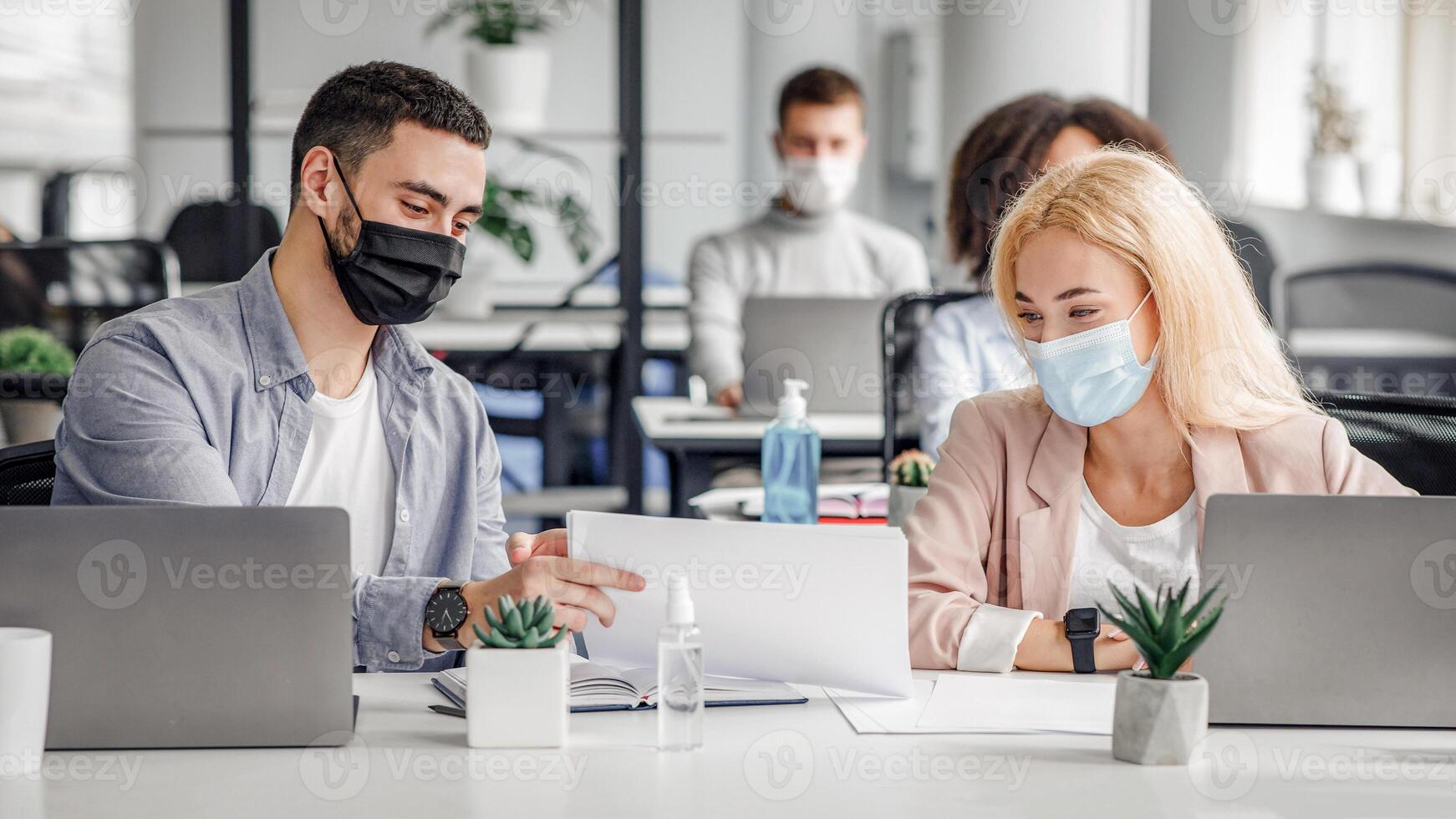 Social distance and work with documents in modern office. Millennial man and woman in protective masks in workplace with laptops and antiseptic, talk after quarantine covid-19, panorama, copy space photo