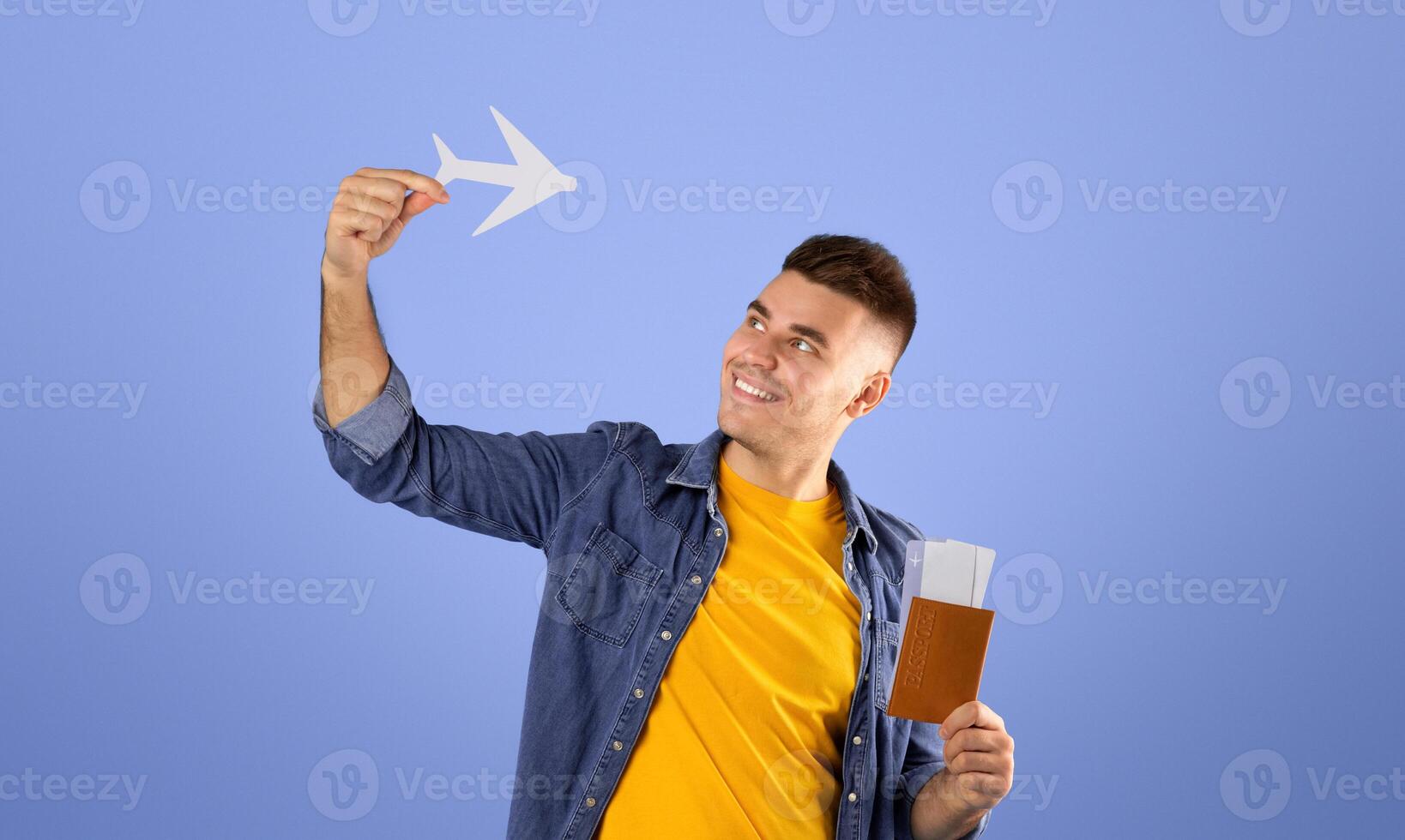 Positive millennial guy with paper plane, tickets and international passport on blue studio background. Young male passanger ready for his abroad vacation. Traveling by air concept photo