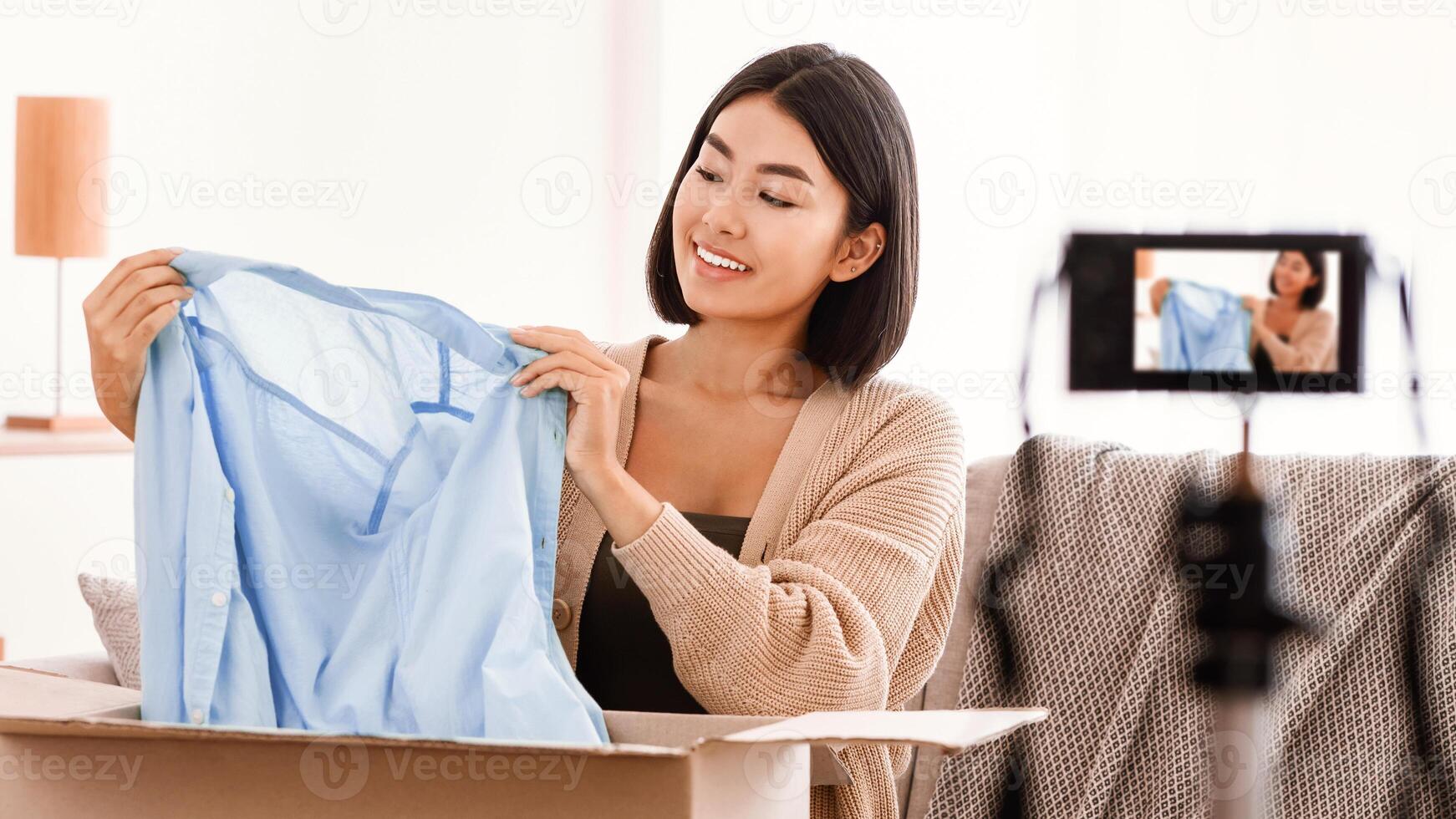 Portrait of happy asian female seller showing stylish blouse on camera on tripod, recording on camera device at home, female vlogger influencer sitting with cardboard box, selling clothes photo