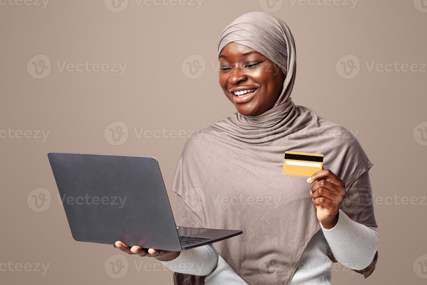 Online Payment. Cheerful Black Woman In Hijab Using Laptop And Credit Card While Standing Over Light Background In Studio, Religious Lady Shopping In Internet Or Making Money Transfer, Copy Space photo