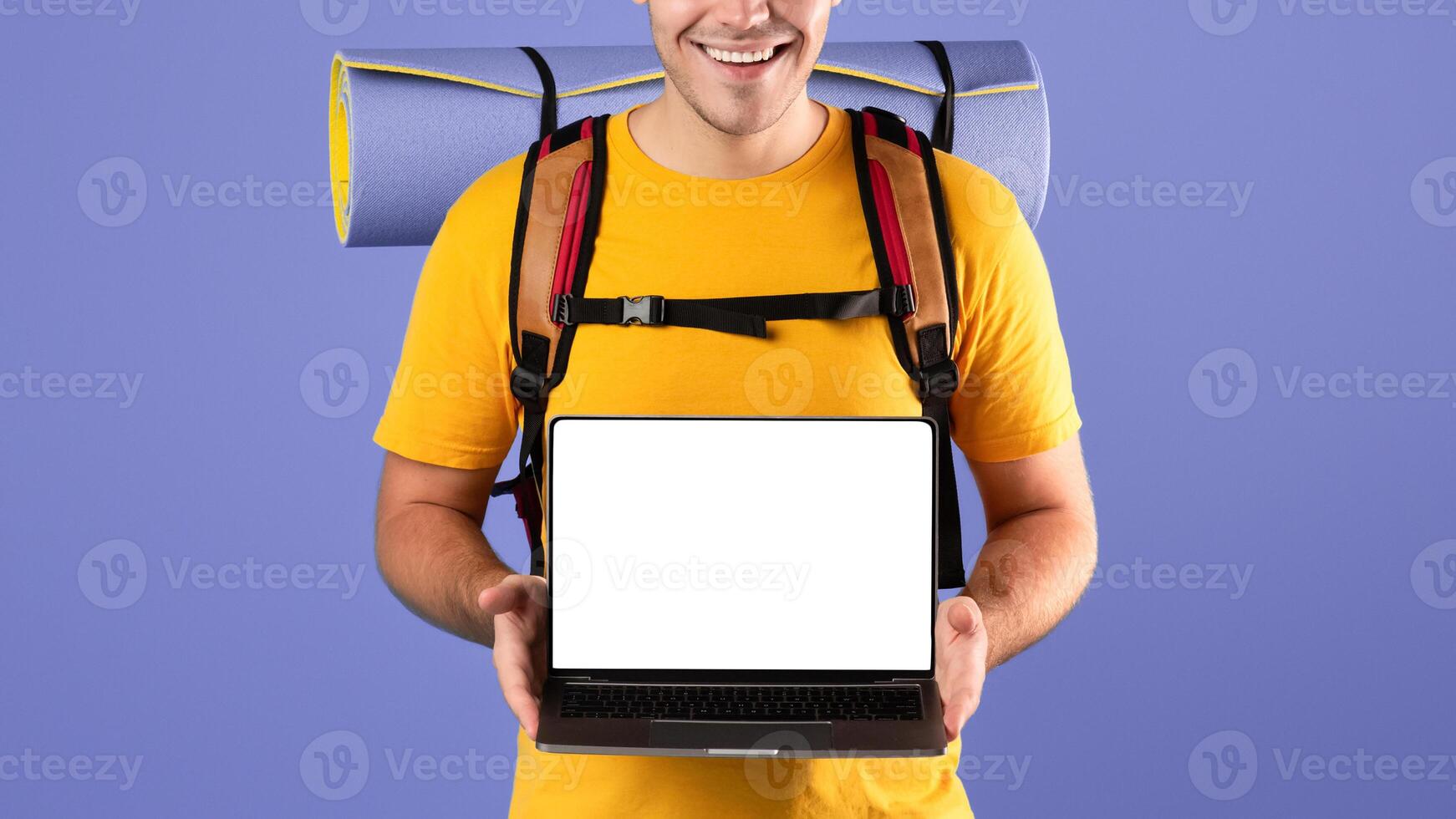 Book your tour online. Positive millennial traveler with backpack showing laptop computer with empty screen on blue studio background. Mockup for tourist agency website photo