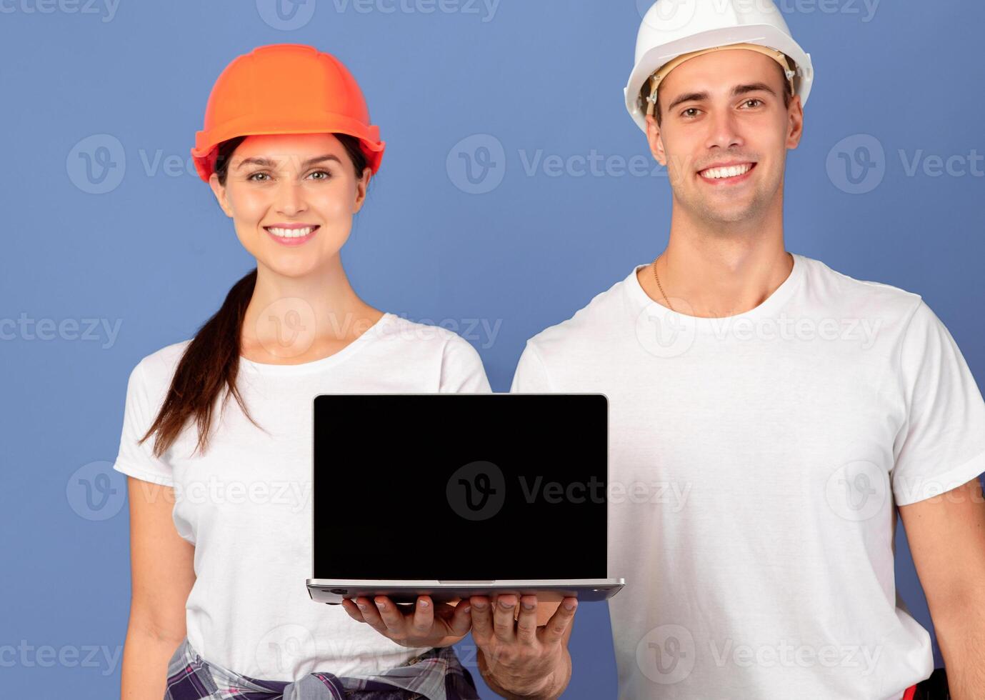 Happy Young Man And Woman In Hardhats Holding Laptop With Black Blank Screen, Demonstrating Copy Space For Design, Recommending Website With Construction Tools, Standing On Blue Background, Mockup photo