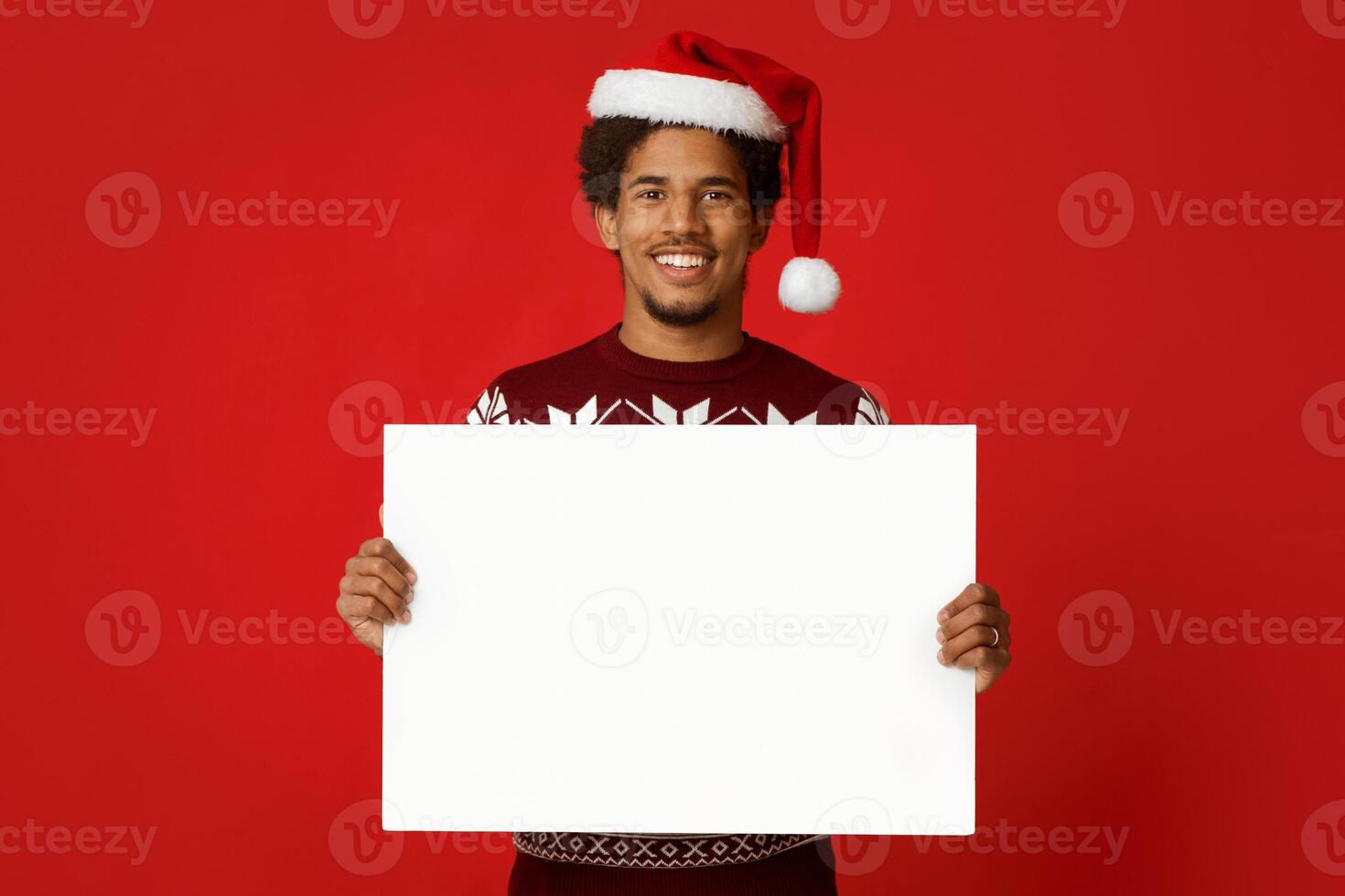 Great offer and advice for new year and christmas. Smiling young african american guy in Santa Claus hat and sweater holds white blank poster and looking at camera, isolated on red background photo