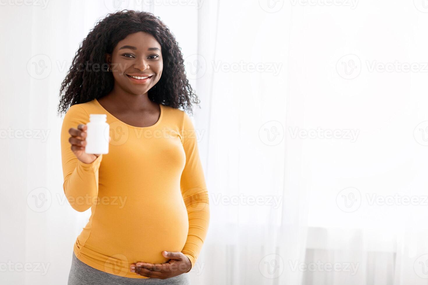 Beautiful pregnant black woman showing her supplements for future mothers, hugging her big tummy and smiling, standing next to window at home, copy space. Nutrition, healthy lifestyle during pregnancy photo