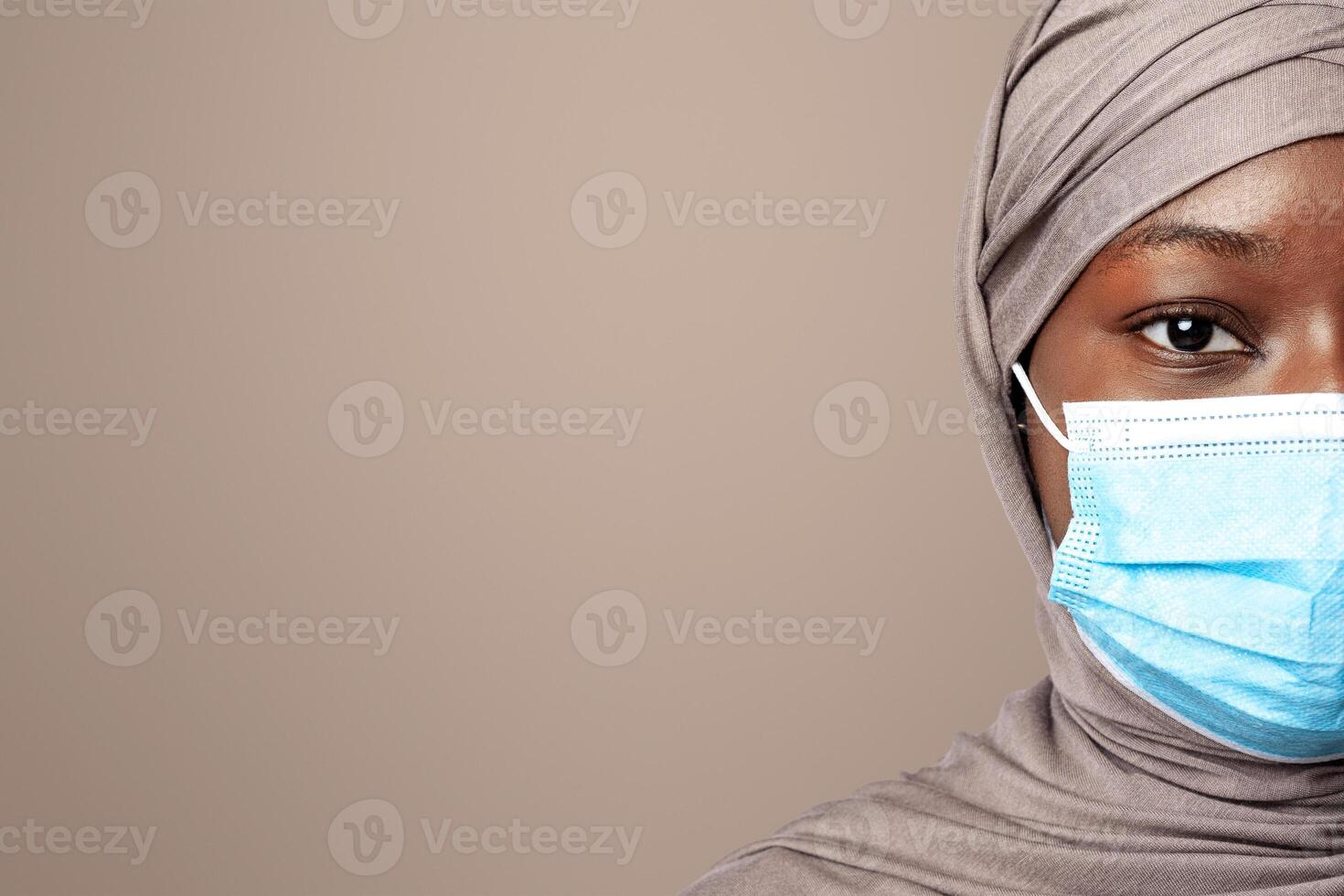 Half Face Portrait Of Serious Black Muslim Lady In Hijab And Protective Medical Mask Posing Over Light Studio Background, Looking At Camera, Follows Coronavirus Preventive Measures, Copy Space photo