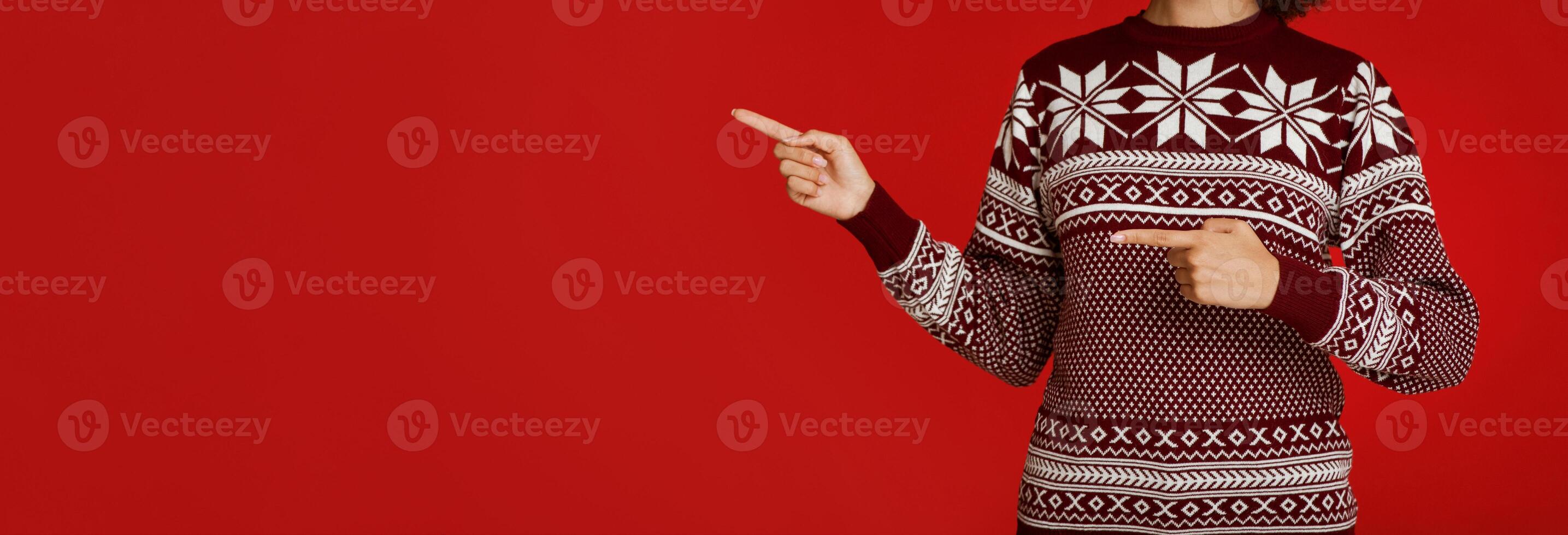 Advice for Christmas and New Year sale at winter holidays. African american lady in sweater with pattern points fingers at empty space, isolated on red background, panorama, cropped, studio shot photo