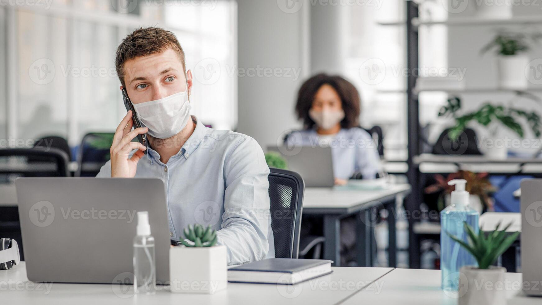 New rules for taking care of health at work after quarantine. Young man in protective mask talking on smartphone with client, at table with laptop and antiseptic in interior of office, free space photo