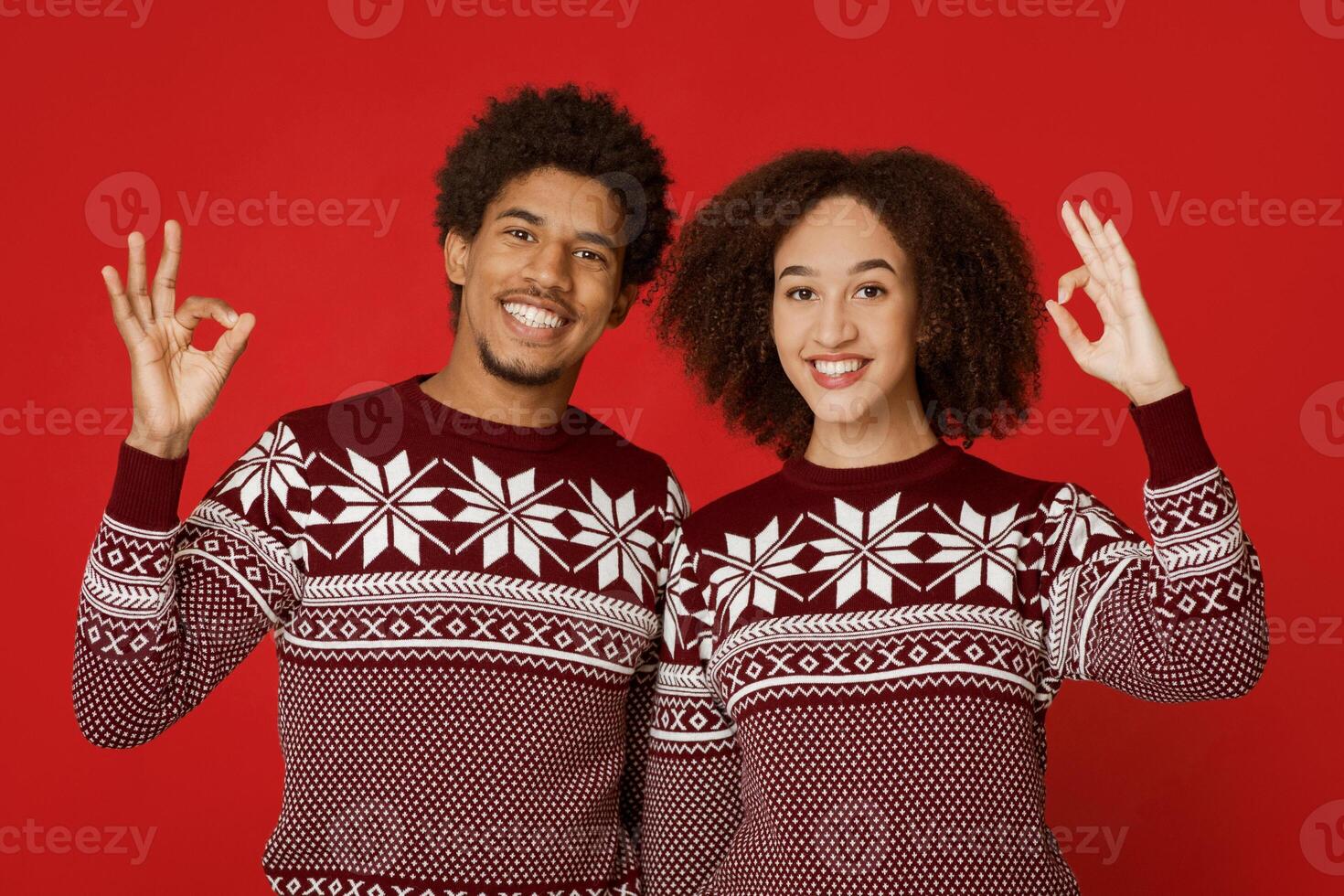 Great New Year and Christmas party. Joyful african american family in identical sweaters show sign okay with their hands and look at camera, isolated on red background, empty space, studio shot photo