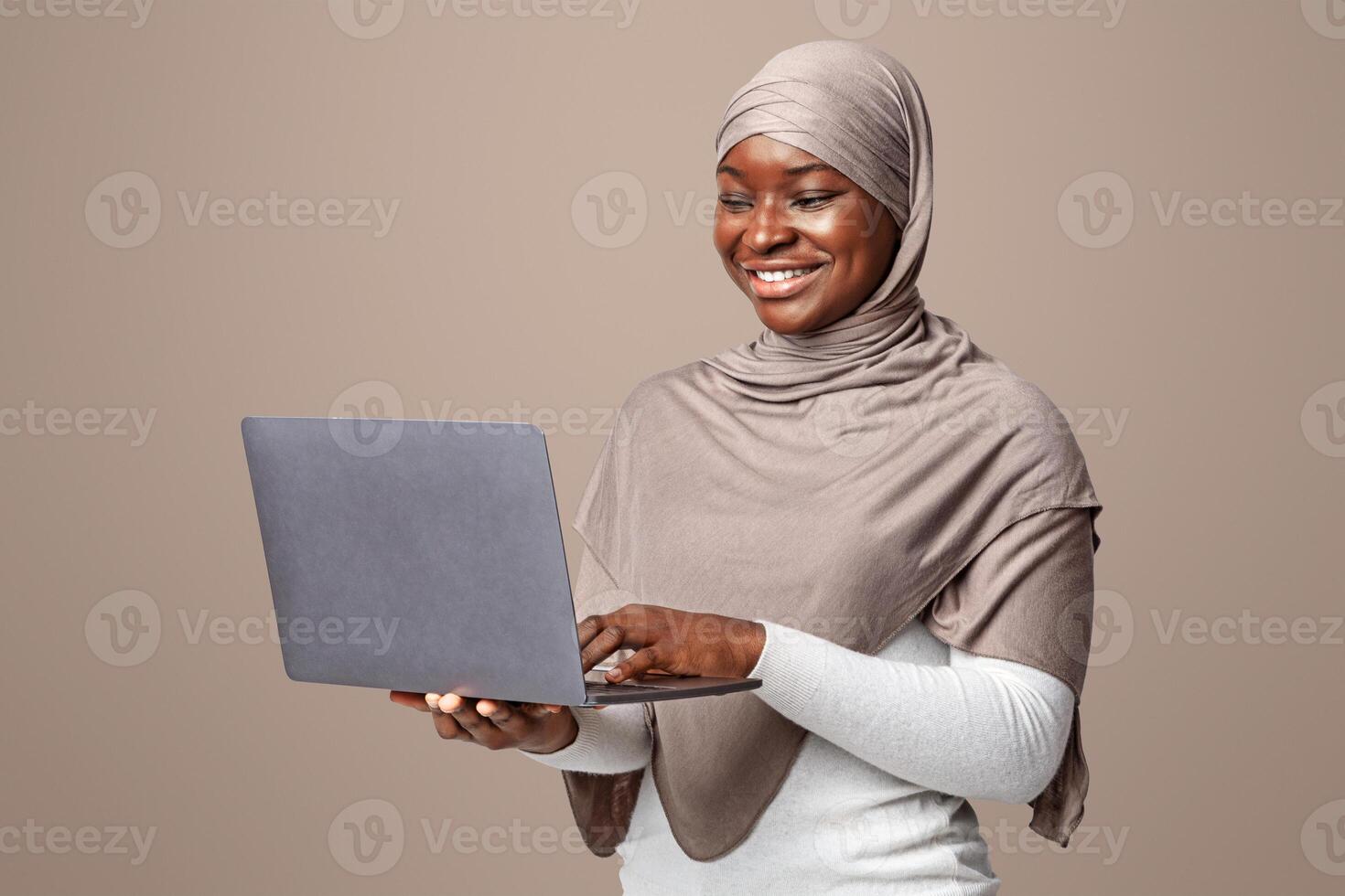 Smiling Black Muslim Lady In Hijab Holding Laptop, Typing On Keyboard. Positive African Islamic Woman Using Computer For Online Shopping Or Remote Work, Standing Over Light Studio Background photo