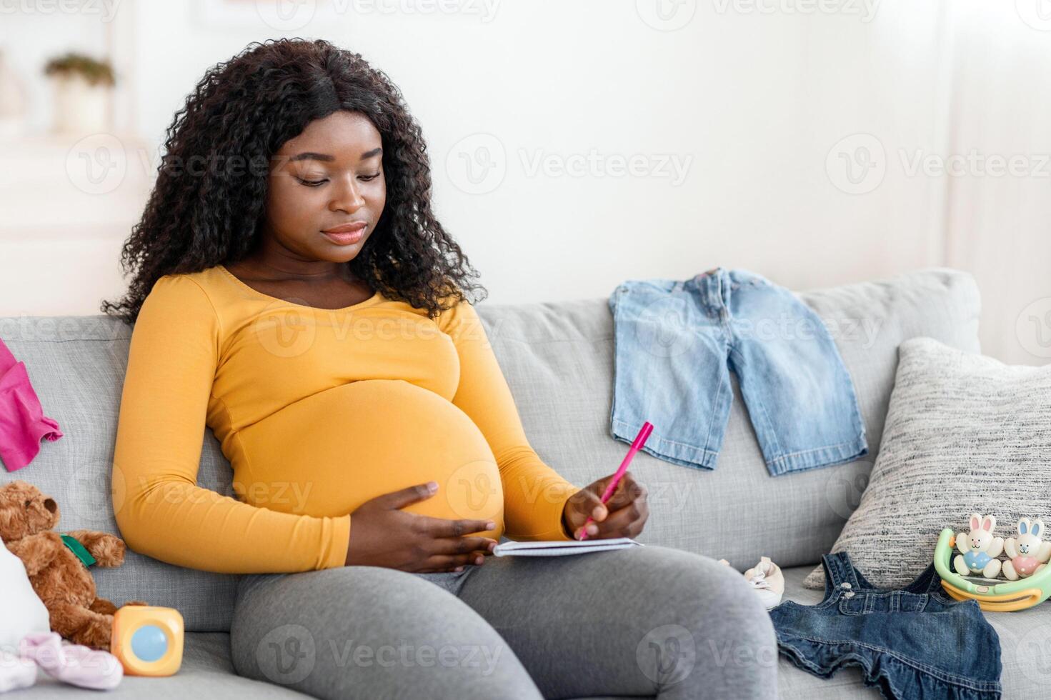 Concentrated pregnant black woman sitting on sofa at home with notebook and pen, making to do list before delivery, free space. Pretty african american single mother getting ready for baby coming photo