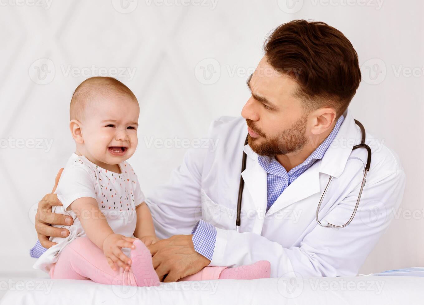 Baby Crying On Medical Checkup With Male Doctor Pediatrician While He Examining Little Toddler Girl Sitting In Clinic Indoors. Sick Child Patient Shouting During Appointment In Hospital photo