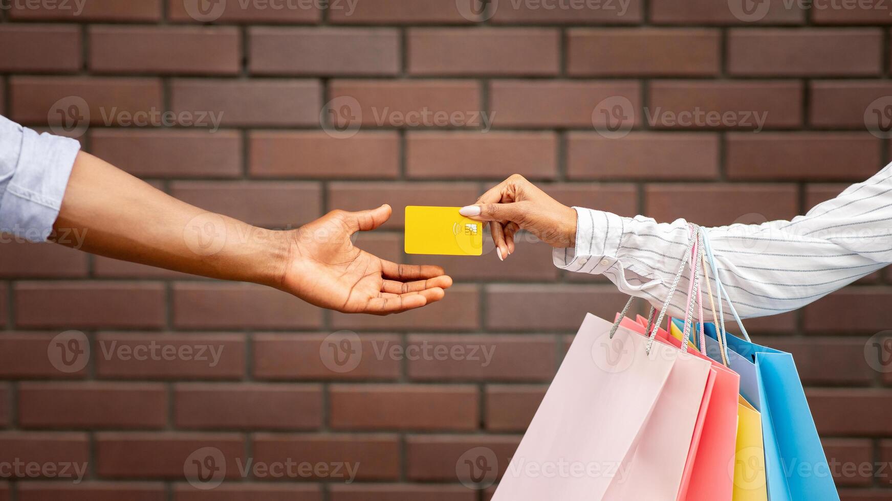 Shopping in boutique. African american girl with multicolored packages gives credit card to seller on brick wall background, cropped, free space photo