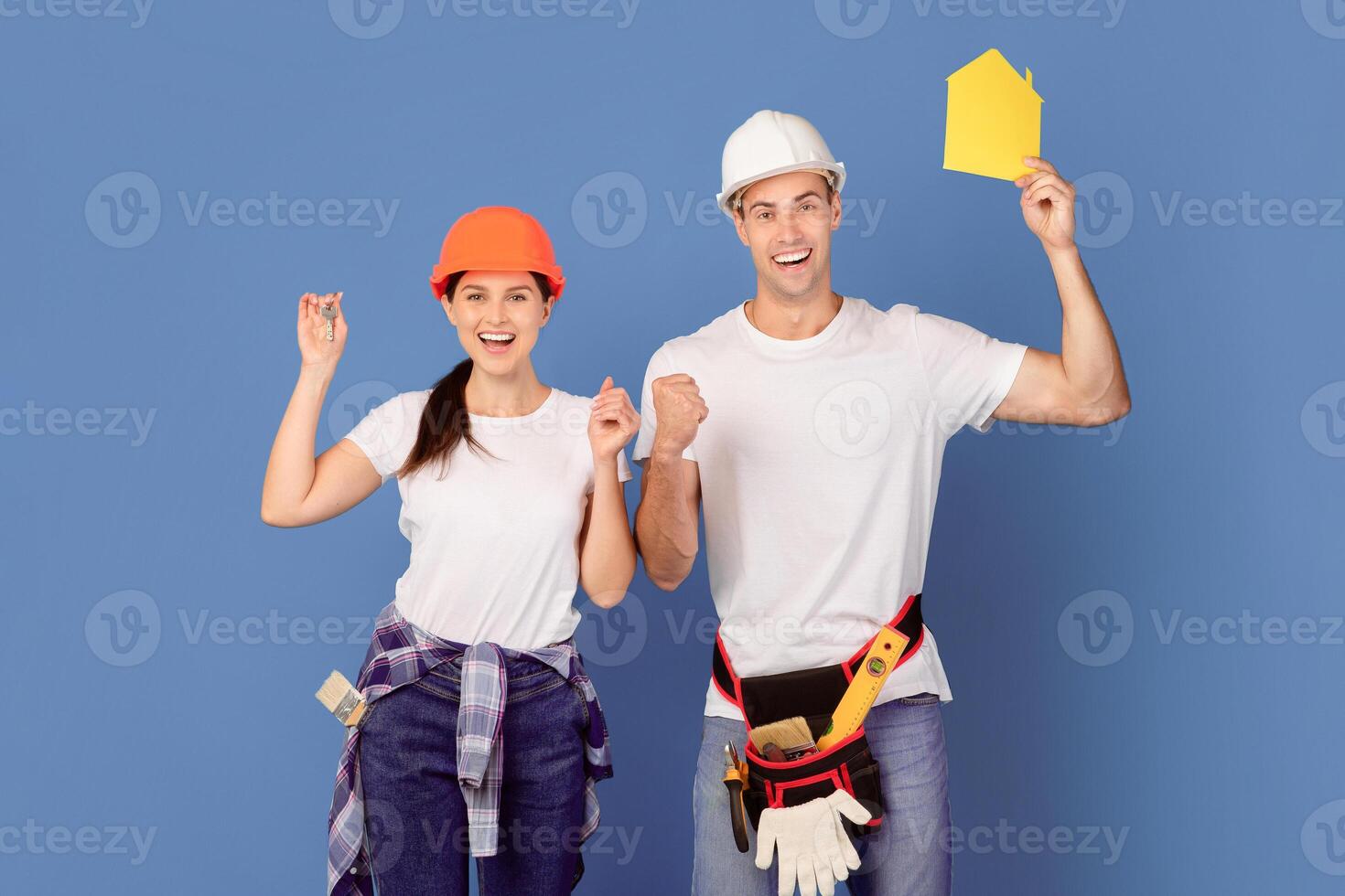 Joyful Millennial Couple In Hard Hats Holding Yellow Paper House Figure And Keys, Celebrating Ending Renovation, Raising Hands In Excitement, Standing Over Blue Studio Background, Copy Space photo