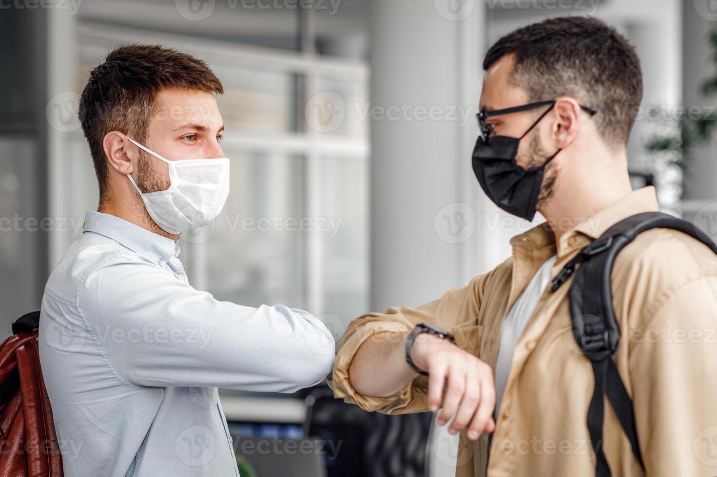 Coworking office and social distance during quarantine covid-19. Millennial guys in protective masks with backpacks greet their elbows, come to work in morning, free space, side view photo