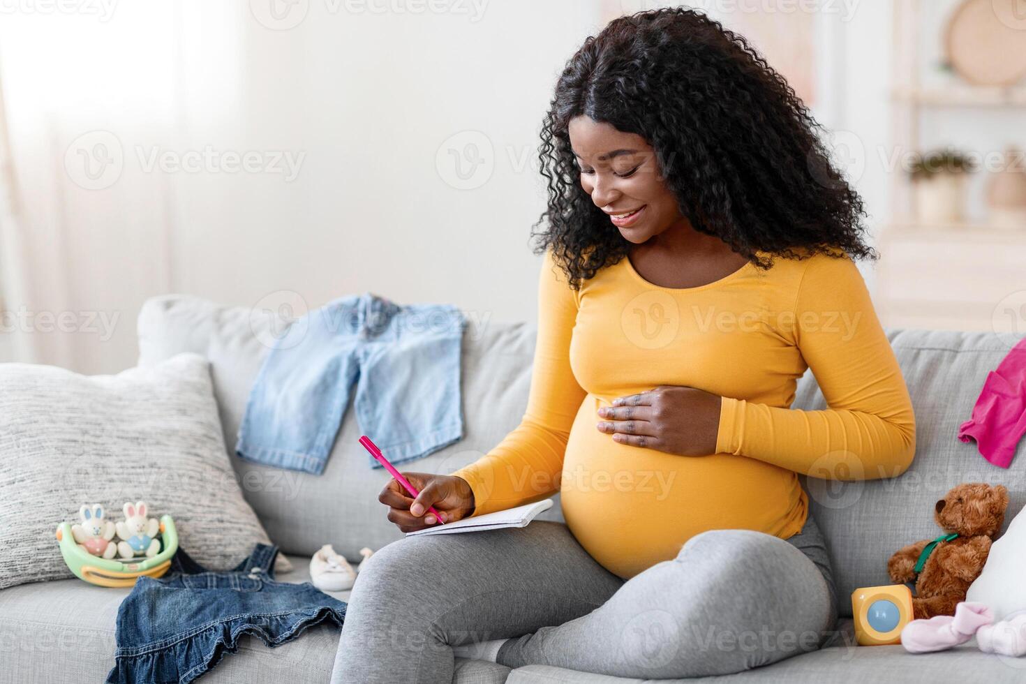 Happy pregnant black woman sitting on sofa with notebook and pen, making shopping list before delivery, free space. Pretty african american single mother getting ready for baby coming, home interior photo