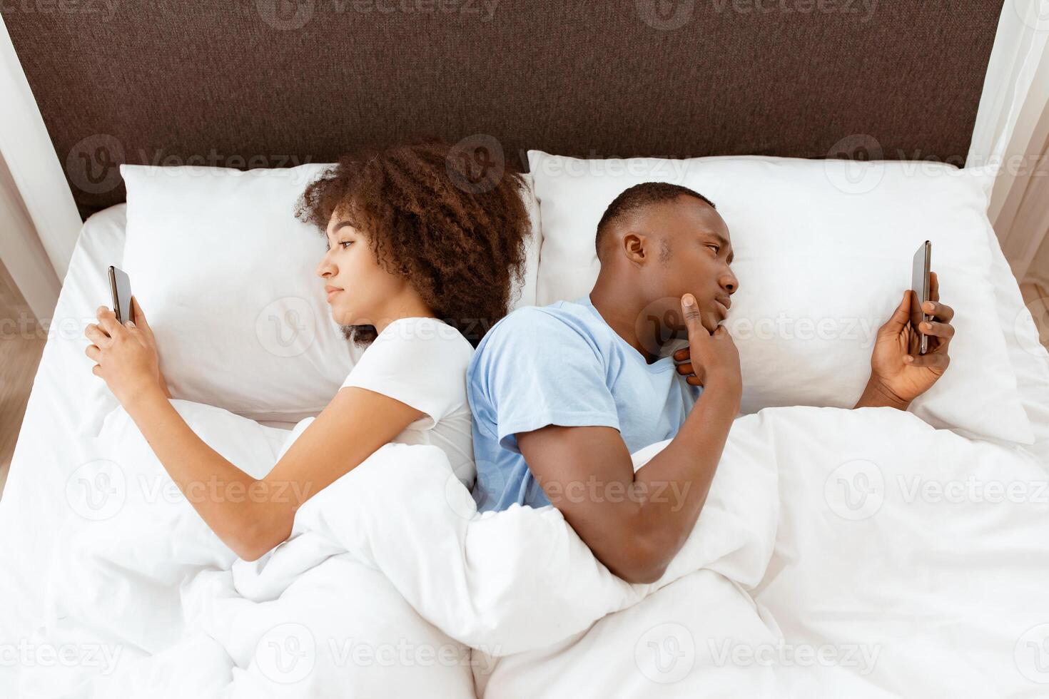 Awake using phone for chat, couple in quarrel, lying on bed back to back, using smartphones. Indifferent, do not pay attention to each other young african american family typing on phones in bedroom photo